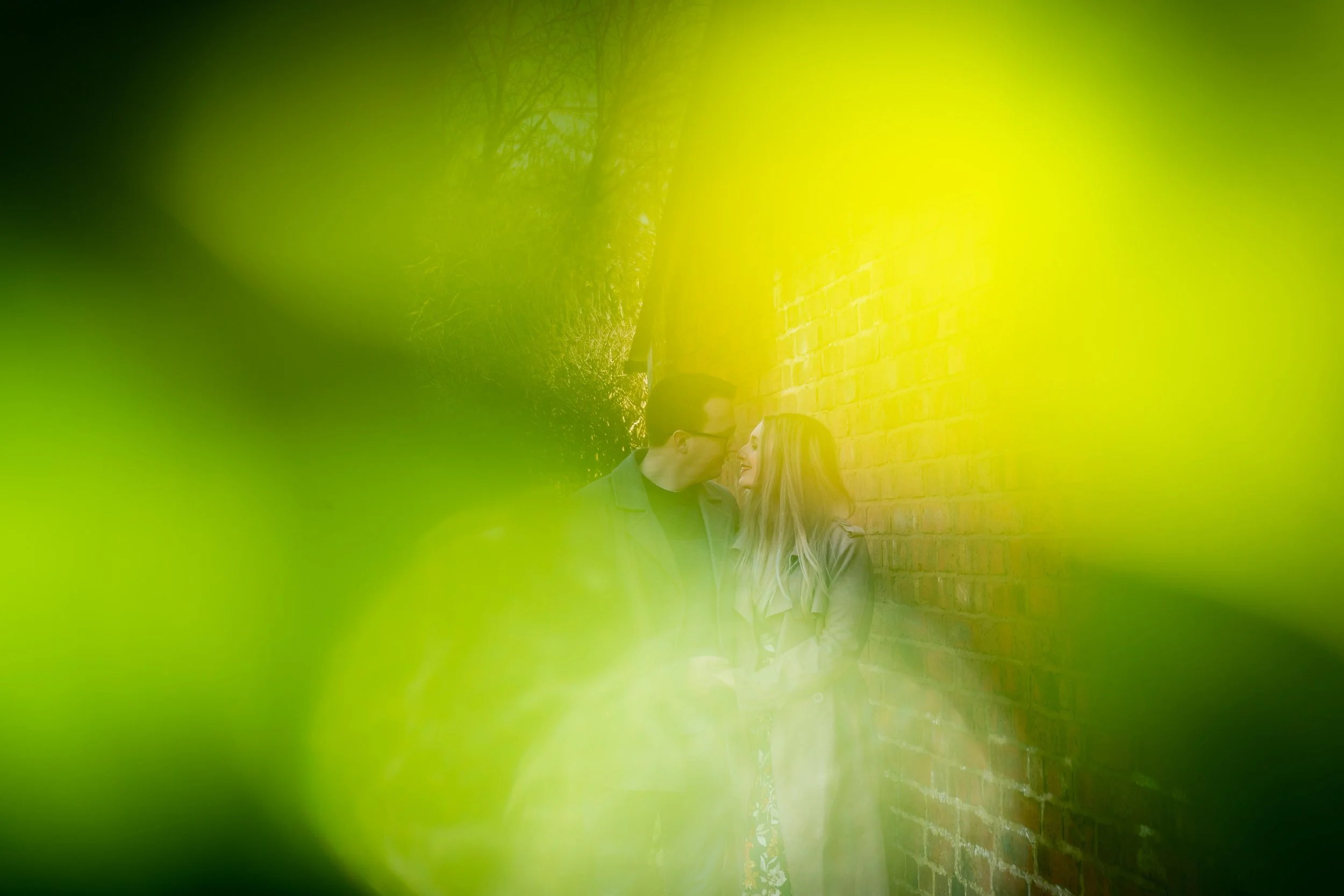 A couple standing close together near a brick wall, viewed through blurred green foliage.