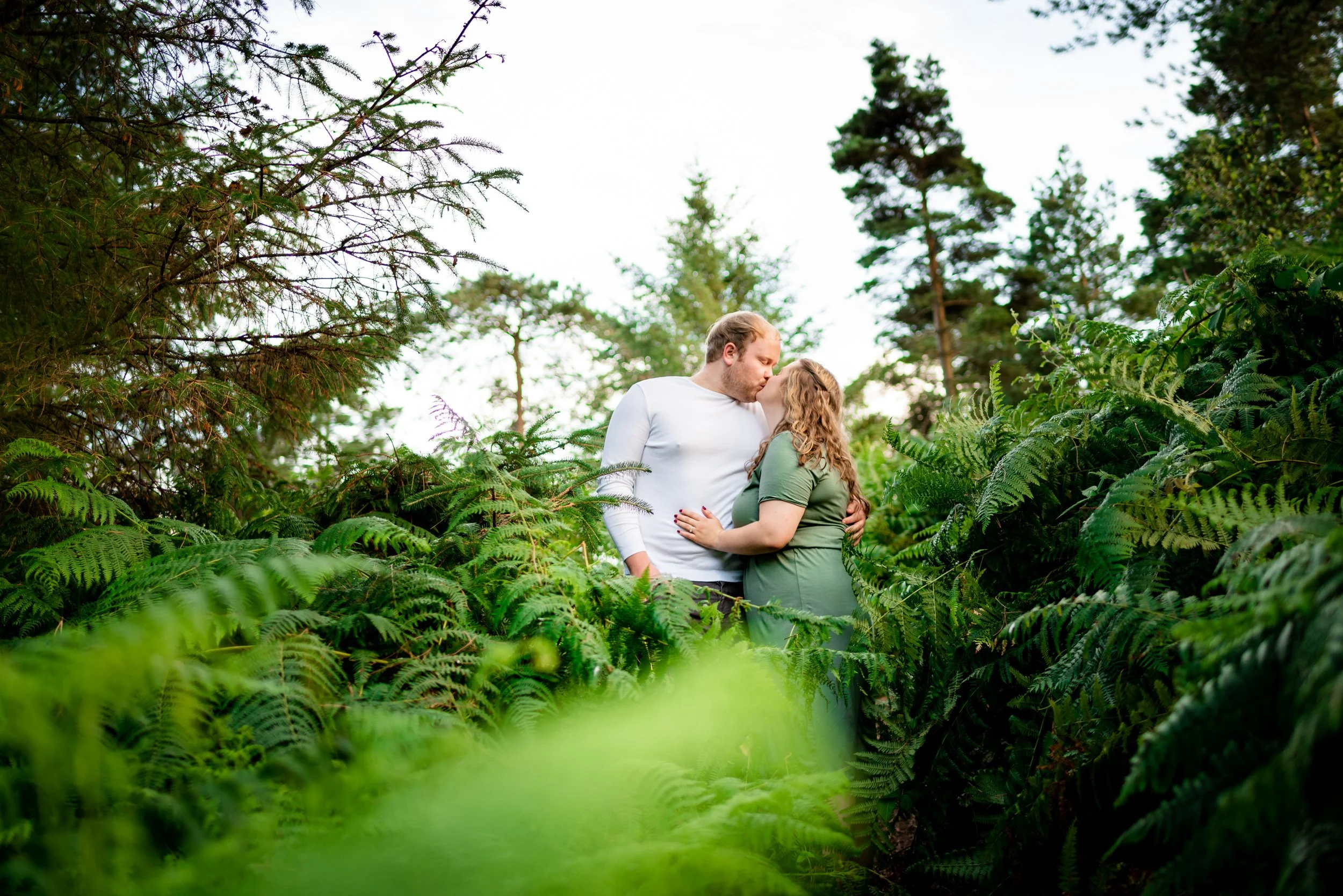 A couple in an embrace, kissing, standing among lush green ferns and trees in a forest or garden setting.
