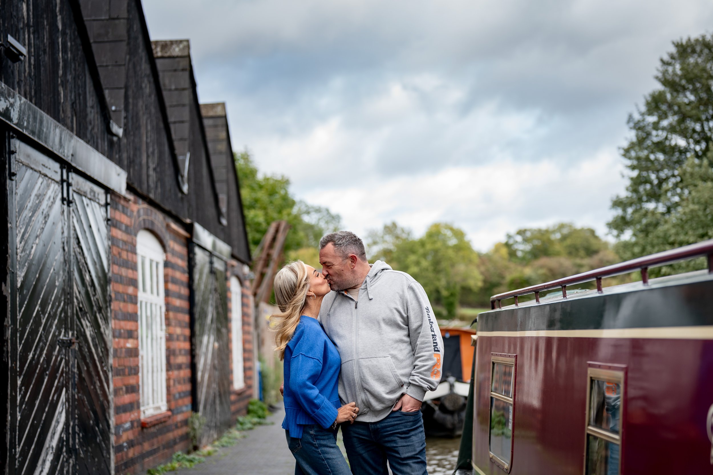 A couple sharing a kiss on a canal path, with a black and red houseboat on the right and a brick building with black wooden siding on the left, under a cloudy sky.