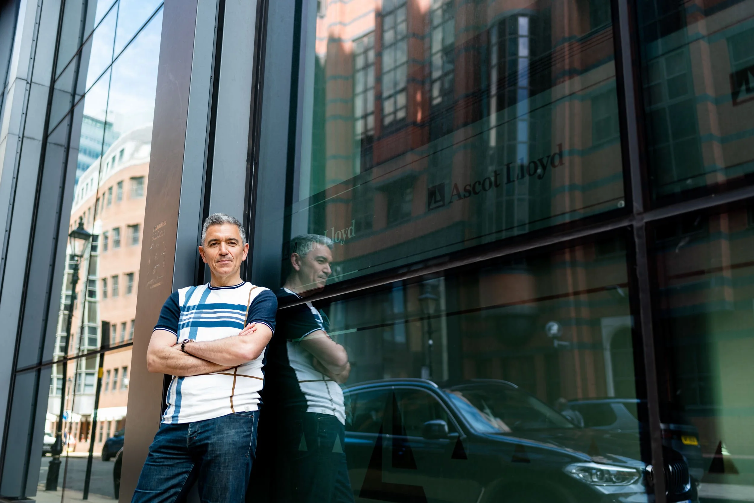 A man standing in front of a reflective glass window on a city street, with his arms crossed. The reflection shows the man and building across the street. The window has the text 'Ascot Lloyd' on it.