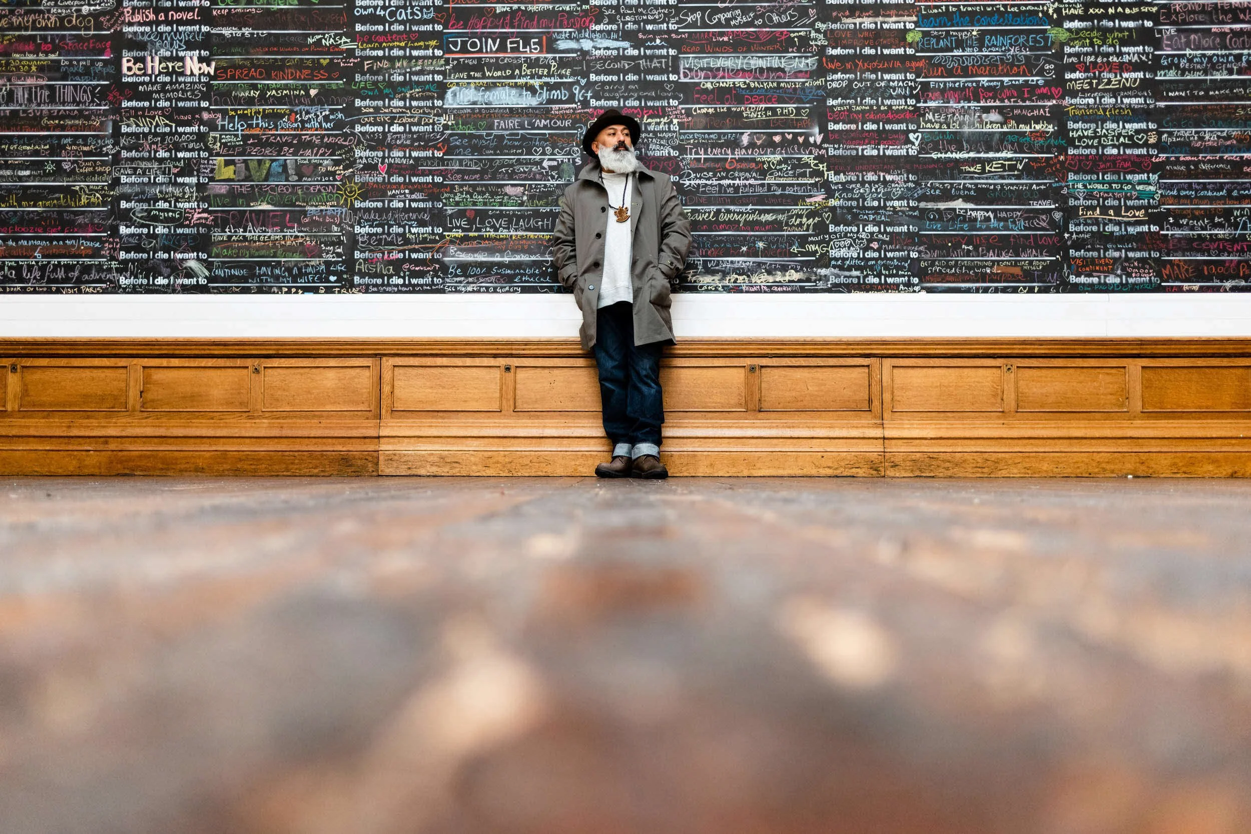 A man with a gray beard and dark hat standing against a colorful chalkboard wall covered in handwritten notes and lists. The wall has a wooden baseboard at the bottom.