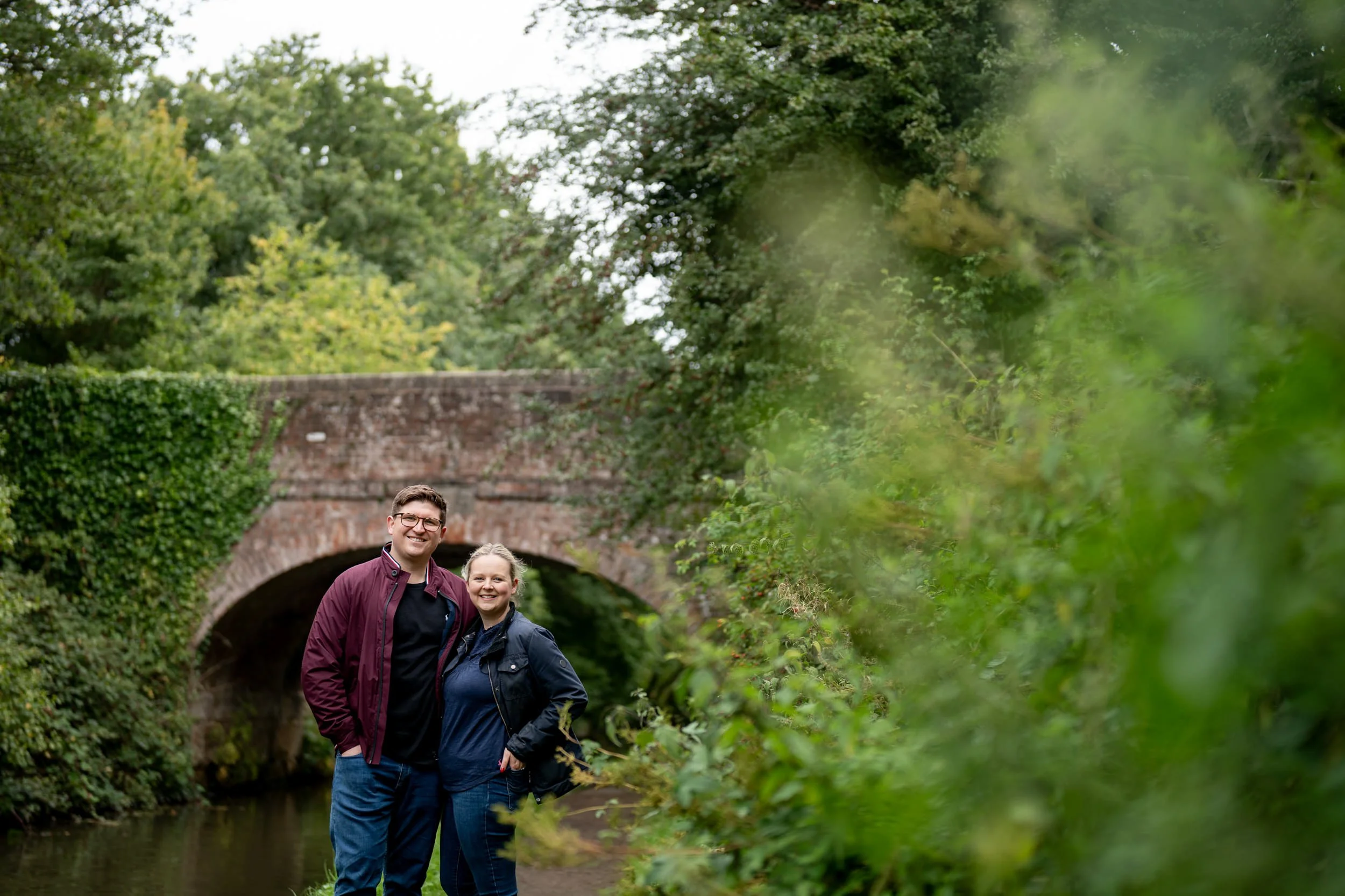 A smiling couple standing by a stream with a stone bridge and lush green trees in the background.