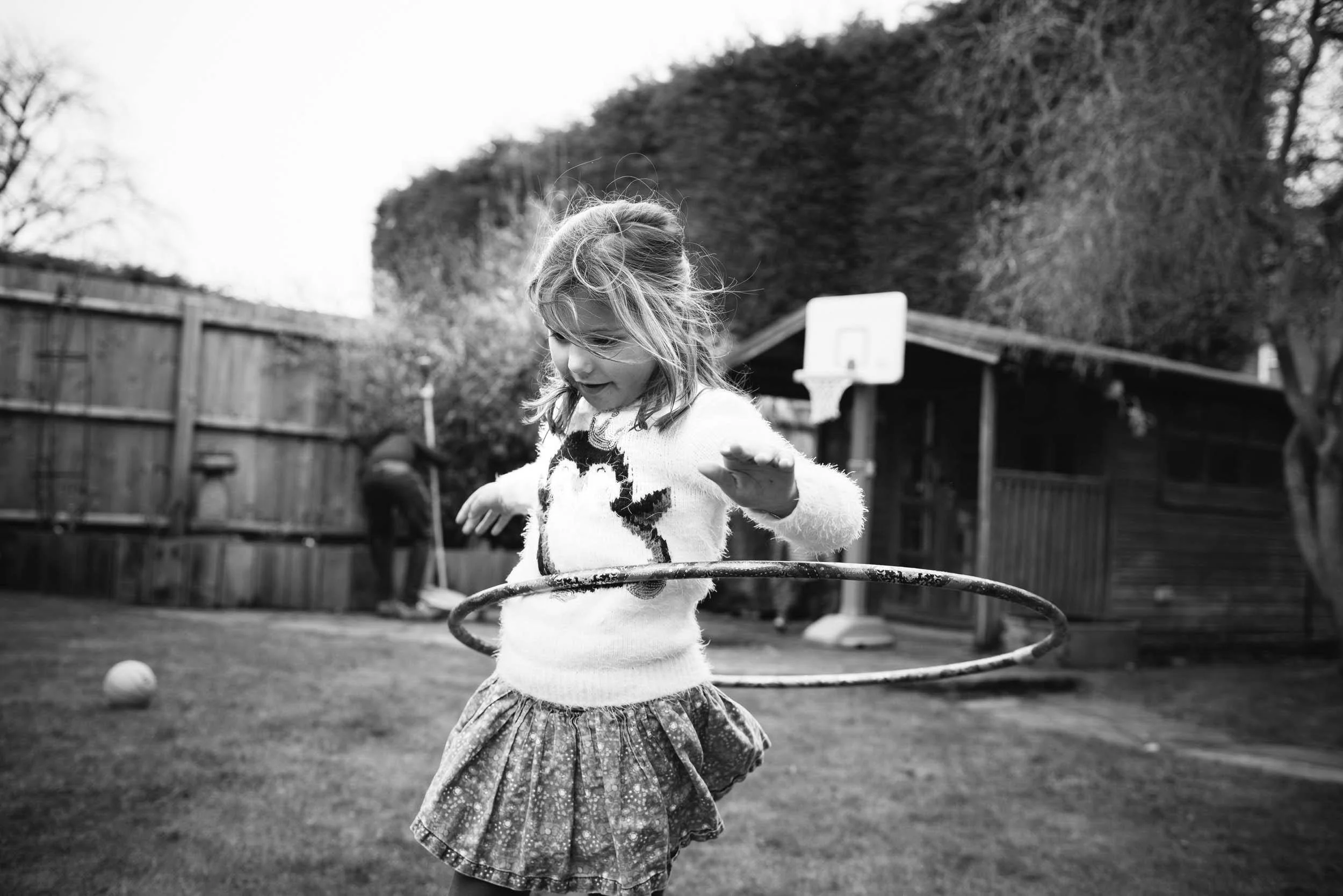 Child playing with a hula hoop outdoors in a backyard with a basketball hoop and wooden fence