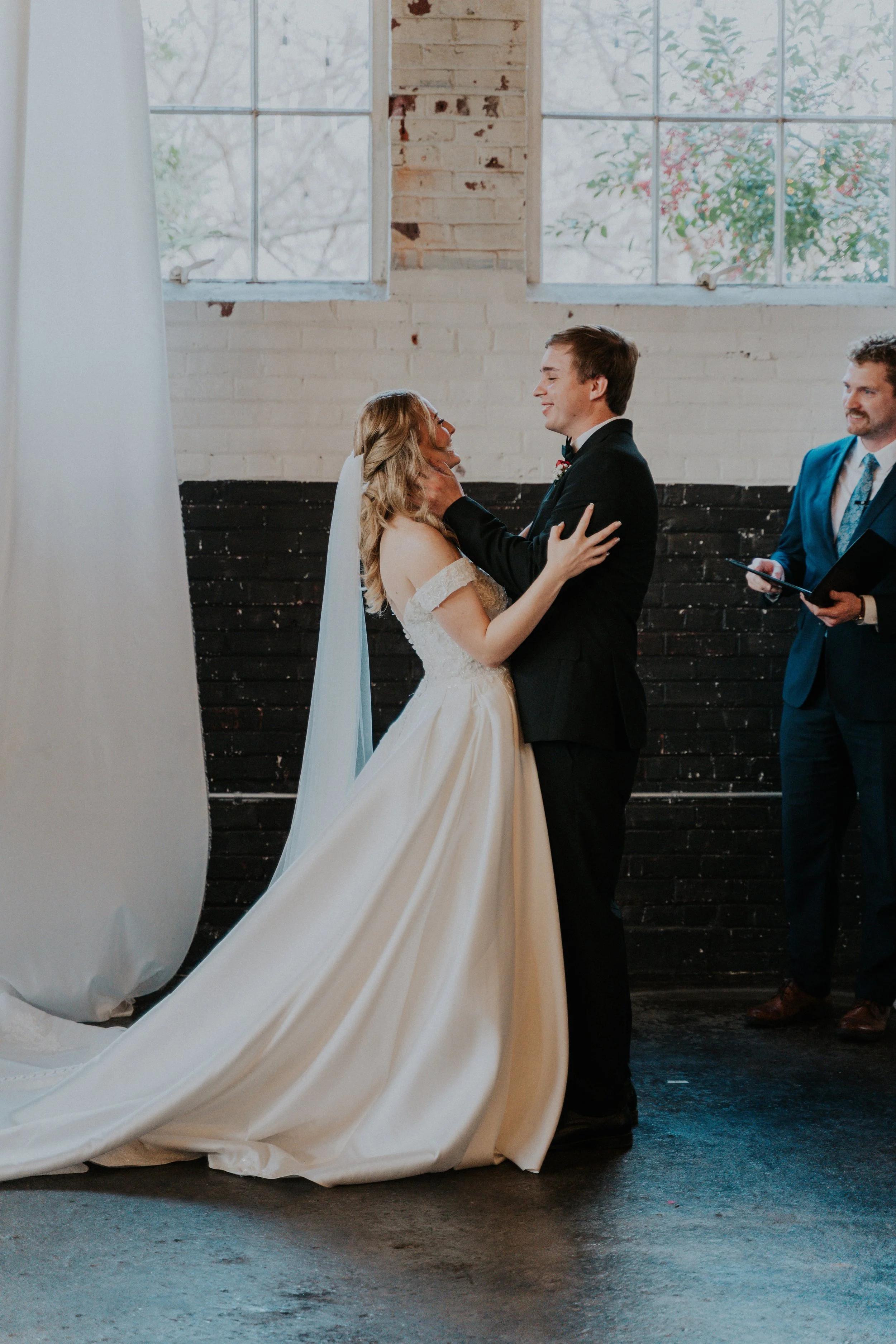 Bride and groom sharing a moment during their wedding ceremony, with an officiant standing to the right, inside a venue with large windows and brick walls.