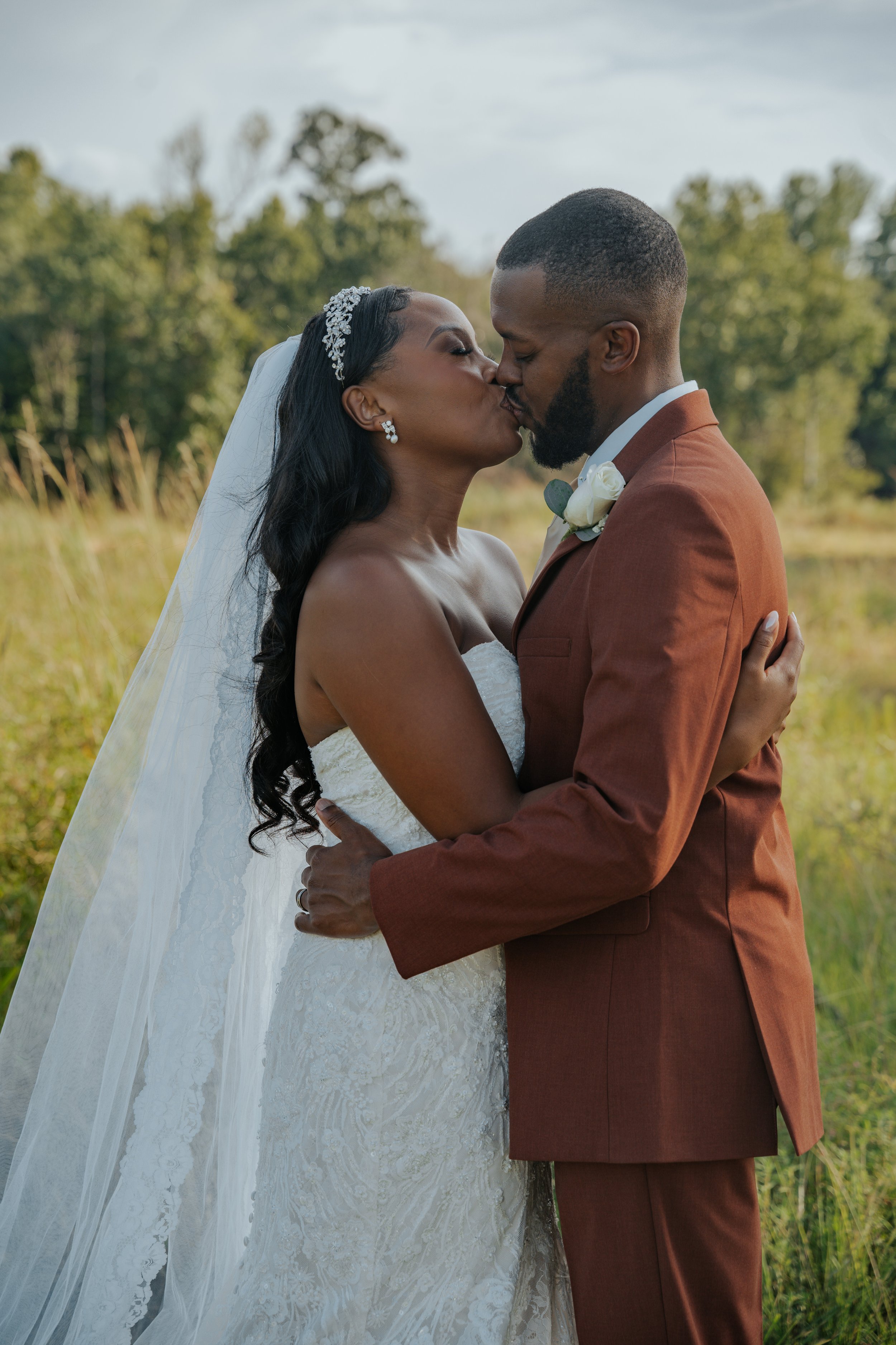A bride and groom kissing outdoors, with trees and grass in the background, on their wedding day.
