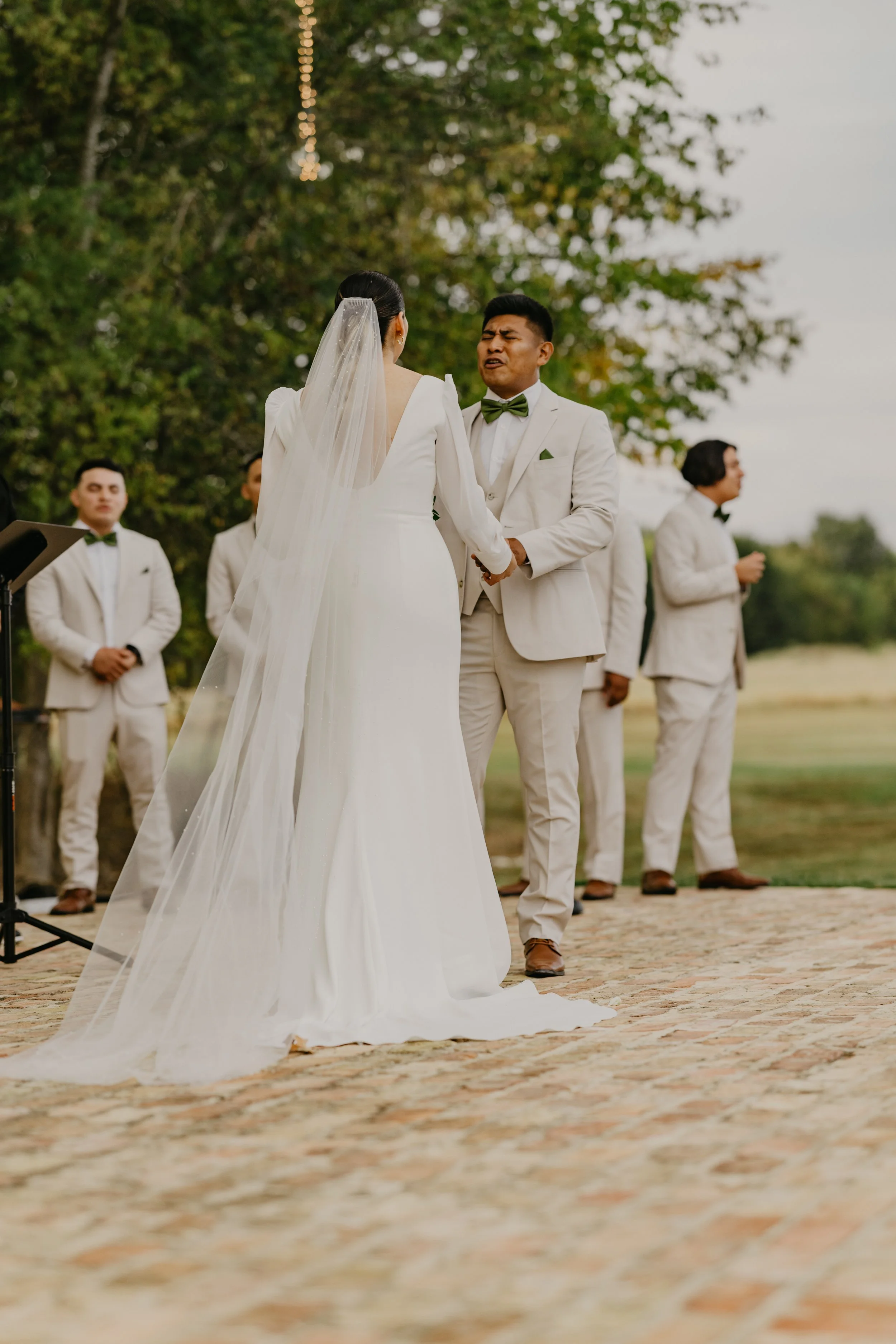 A wedding ceremony outdoors with a bride and groom holding hands and singing, surrounded by groomsmen.