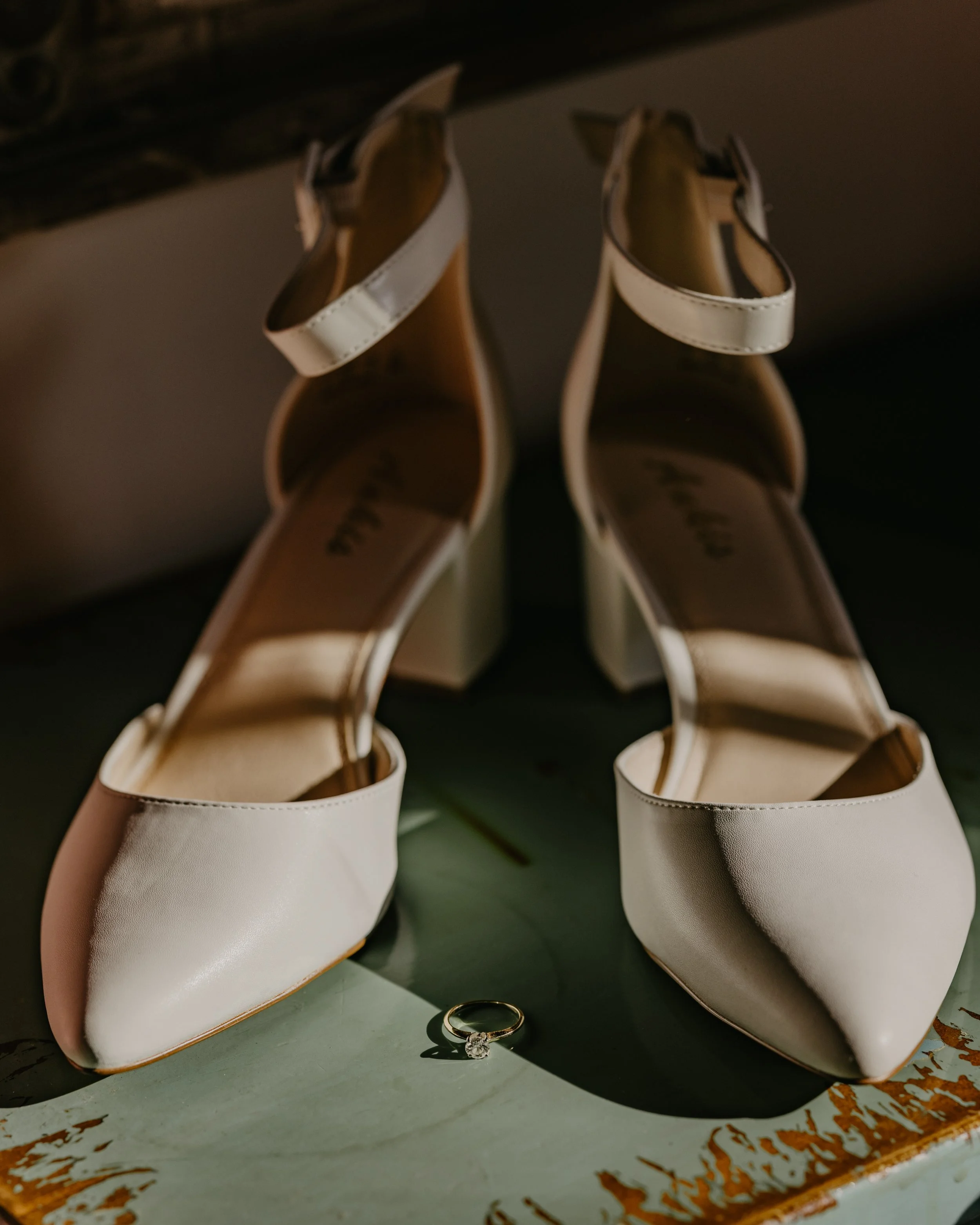 A pair of white high-heeled shoes with ankle straps and a diamond ring placed on a green surface.
