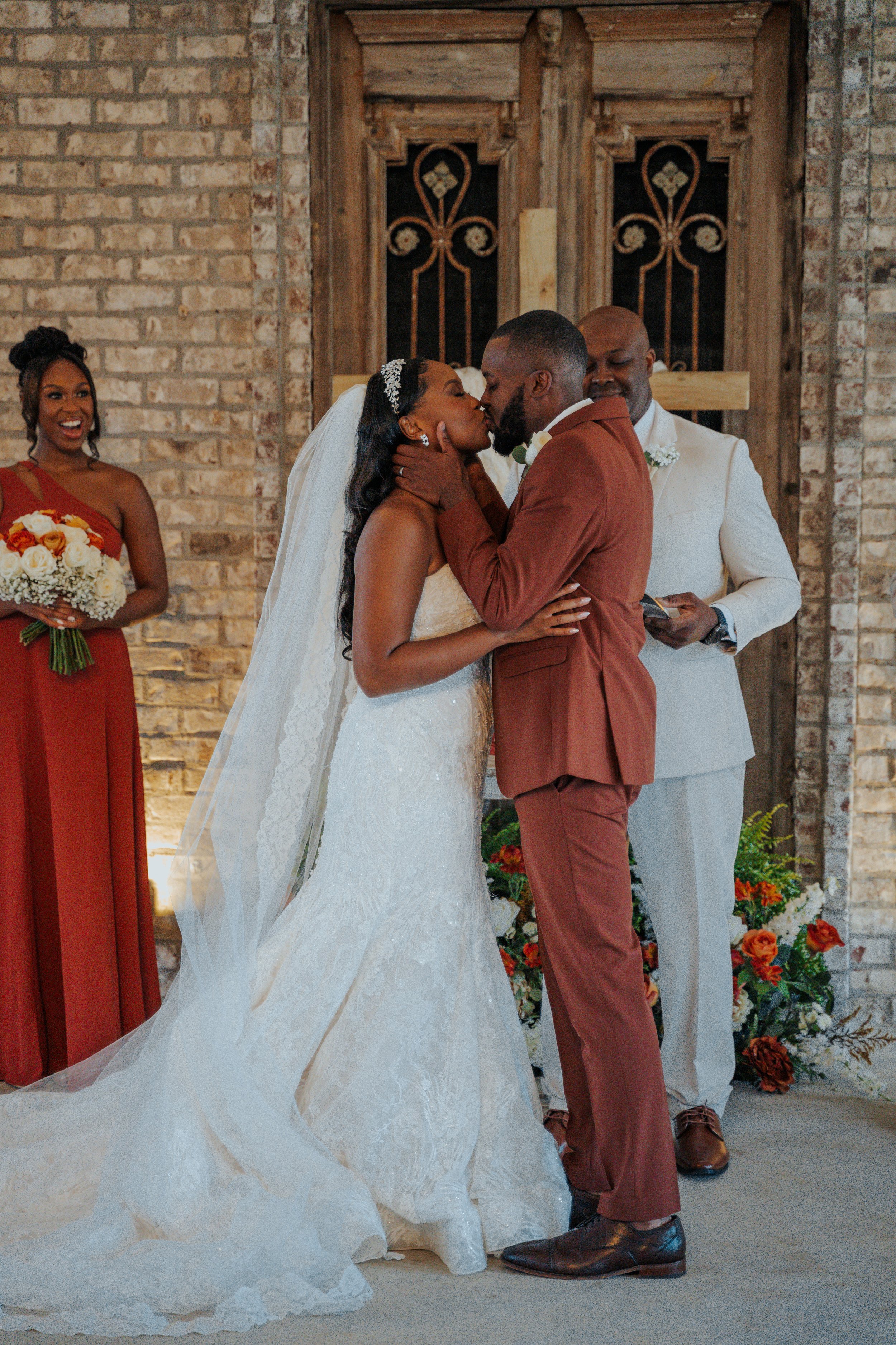 Bride and groom kissing at their wedding ceremony, with a bridesmaid holding a bouquet in the background and a man officiating, in a rustic brick-walled chapel with floral decorations.