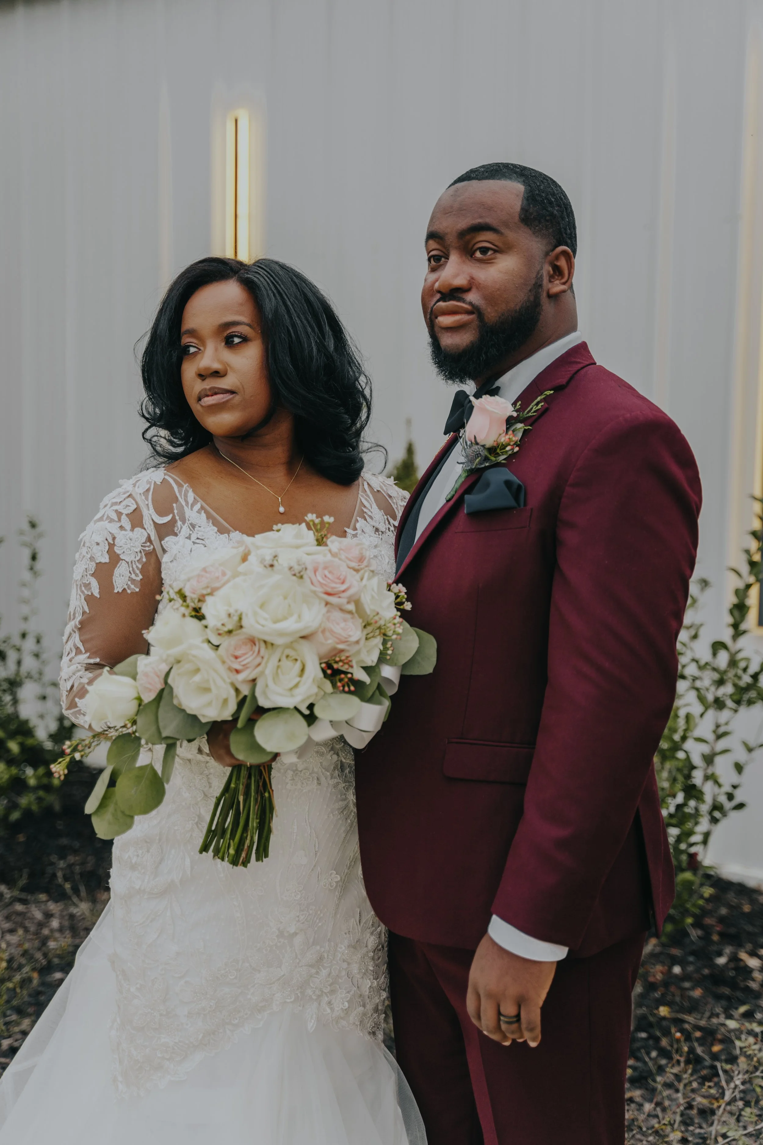 A bride and groom standing together at their wedding indoors, with the bride holding a bouquet of white and pink roses.