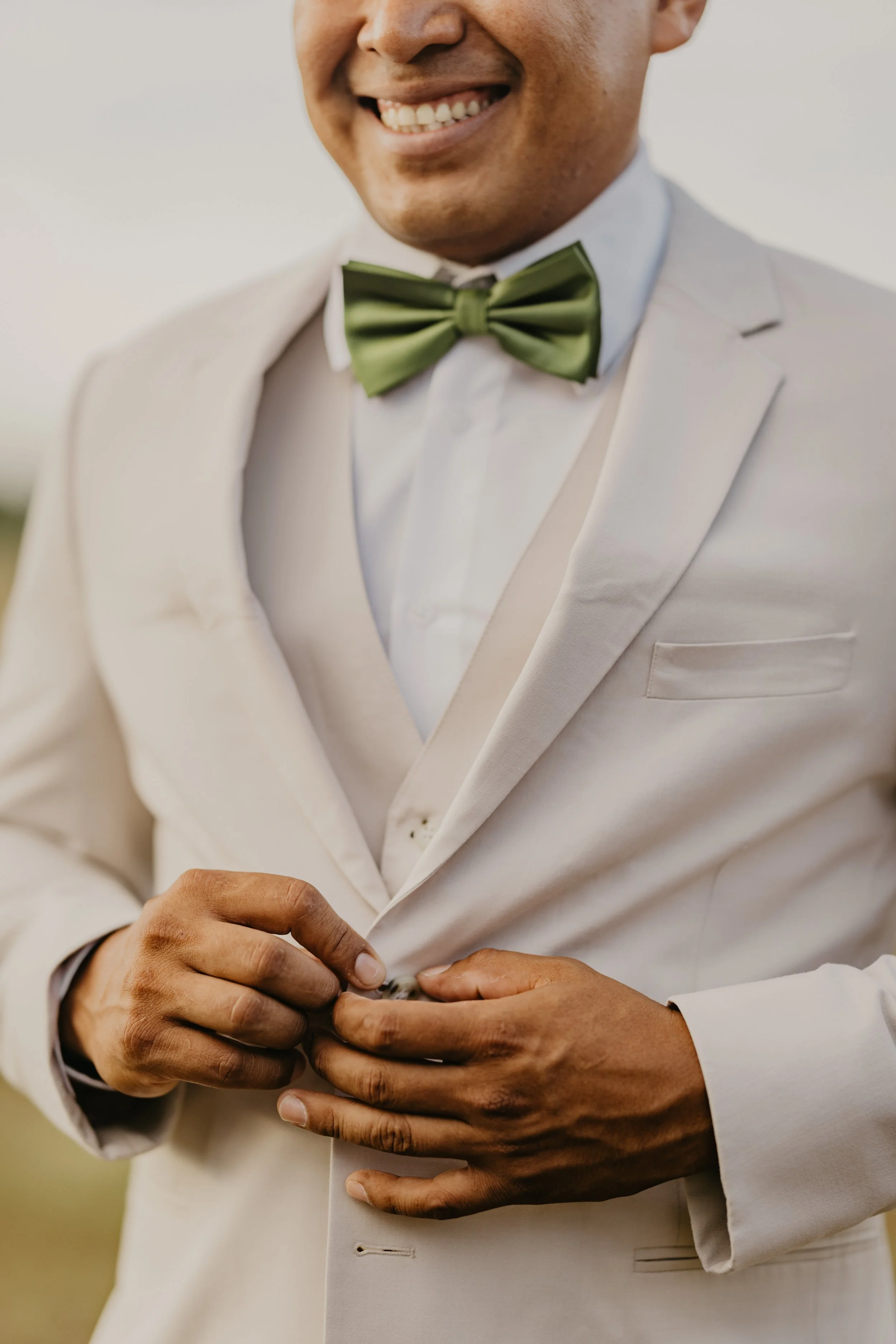 A man smiling, wearing a cream-colored suit and a green bow tie, buttoning his jacket.