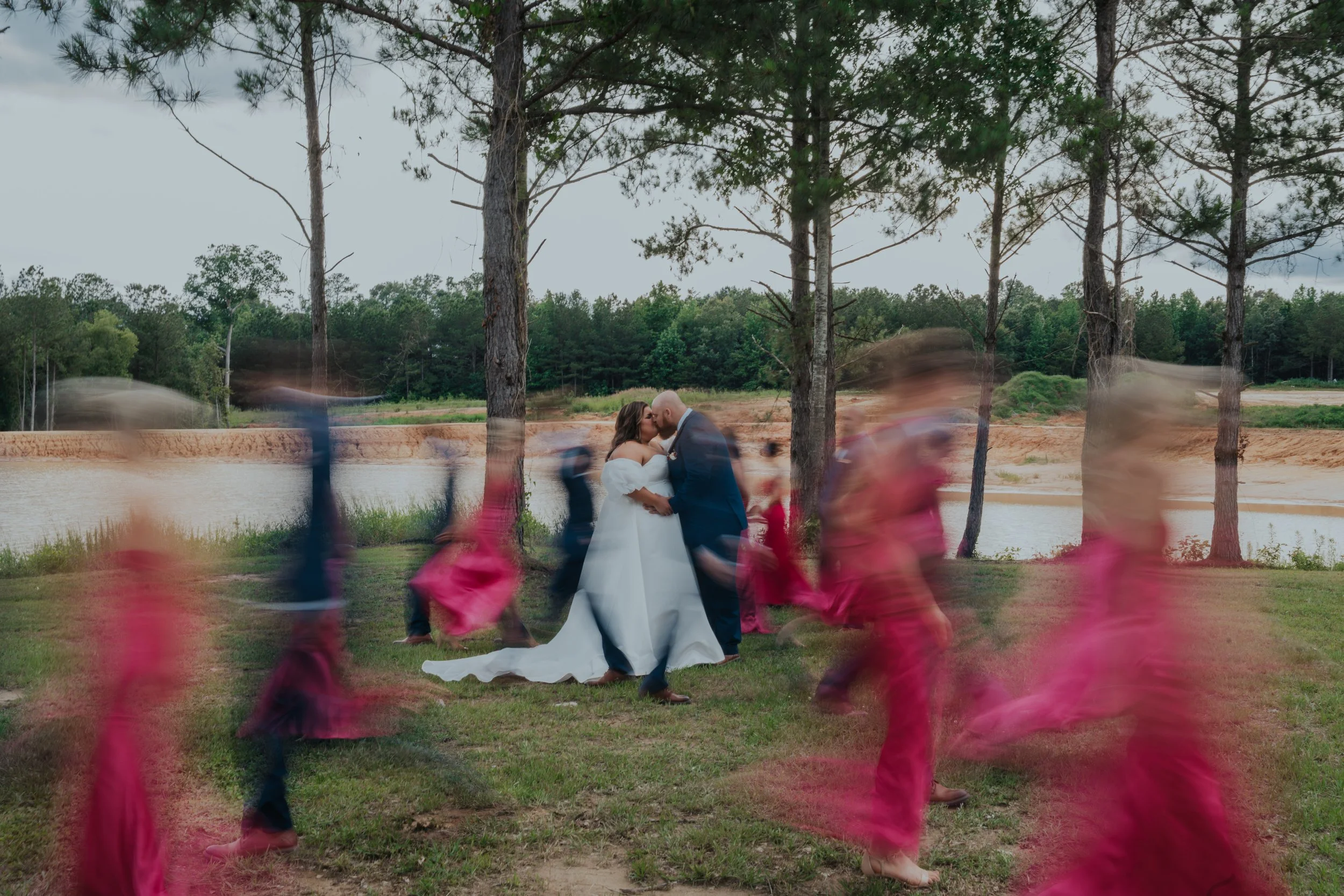 Bride and groom sharing a kiss surrounded by children in pink dresses, with a lake and trees in the background.