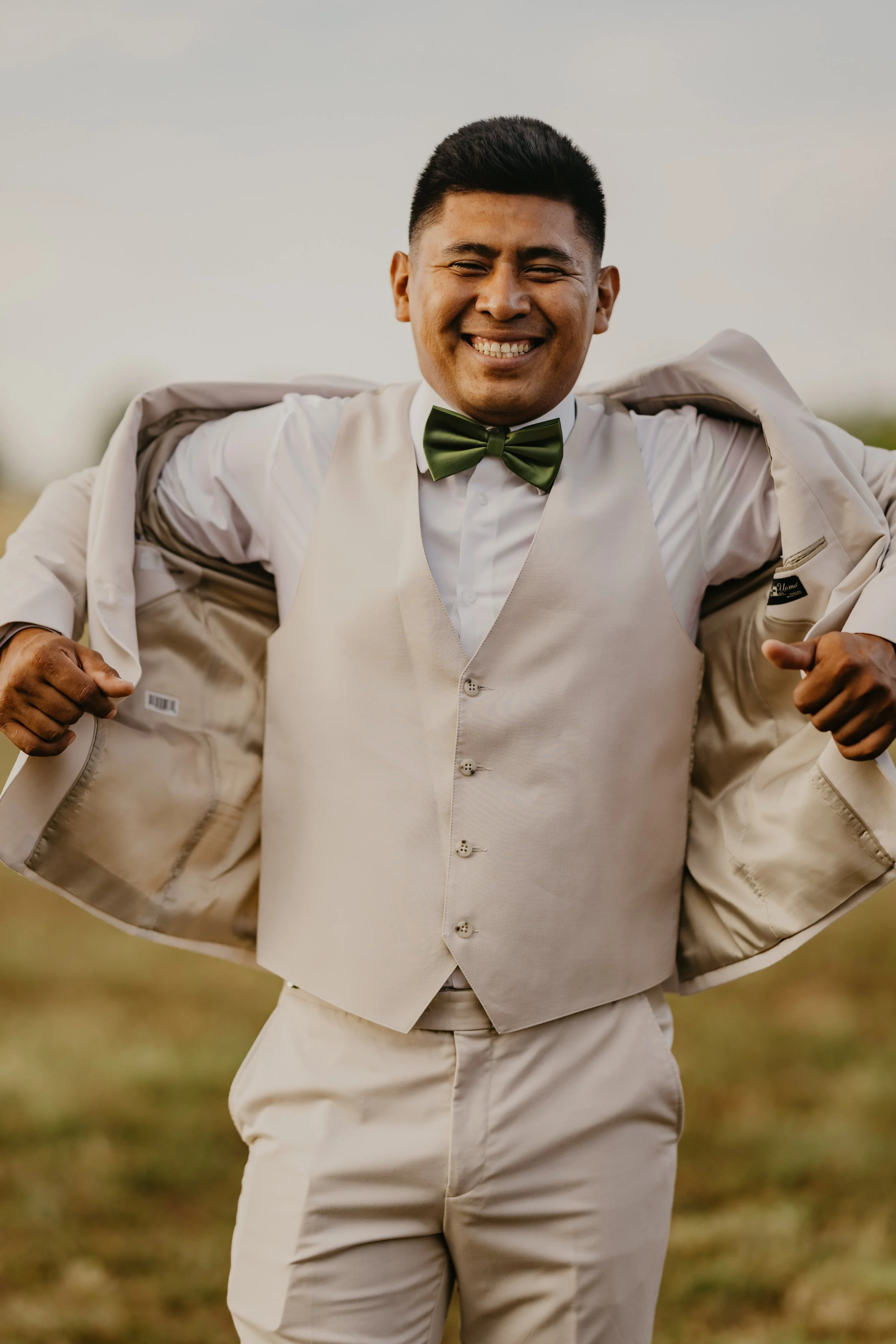 A man dressed in a tan suit and vest, with a white shirt and a green bow tie, is smiling and putting on a matching tan suit jacket outdoors.