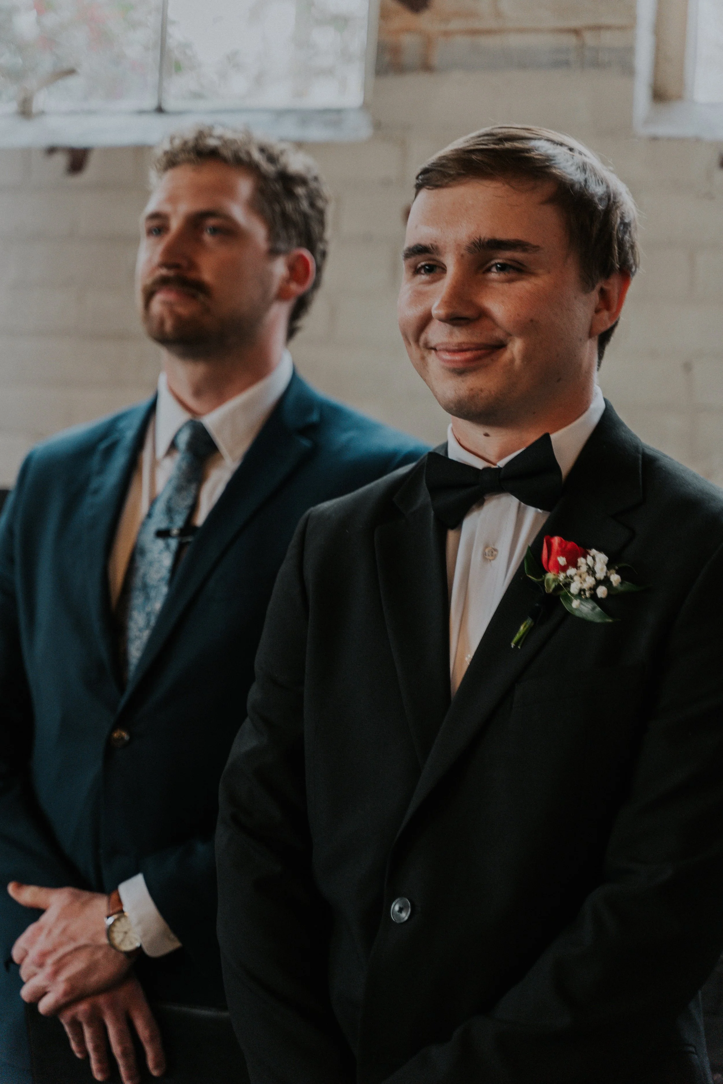 A groom in a tuxedo with a boutonniere standing next to a man in a suit, at a wedding ceremony. The groom is smiling, and sunlight is coming through windows behind them.