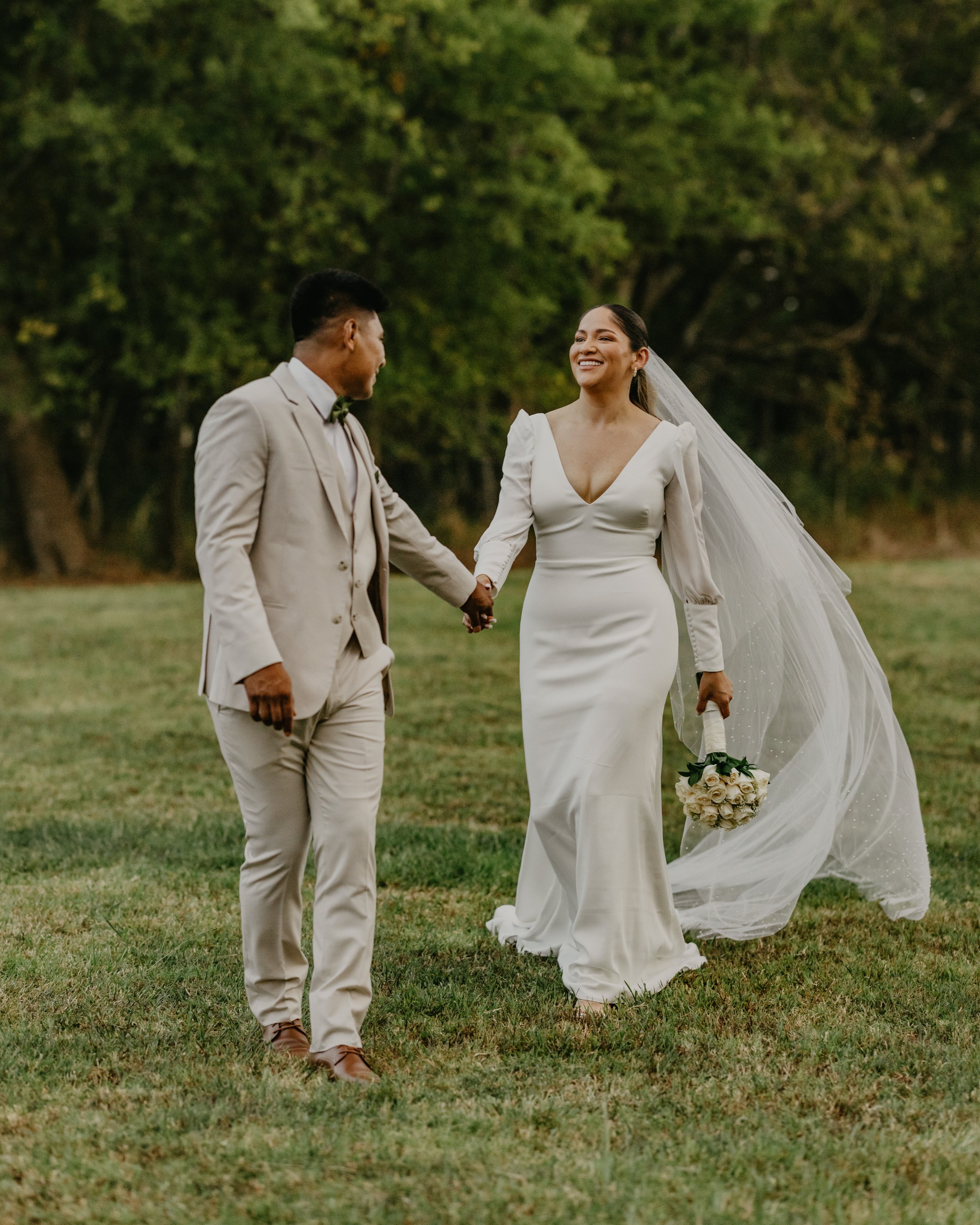 Bride and groom holding hands and smiling outdoors on their wedding day, with the bride wearing a white gown and veil, holding a bouquet of white roses, and the groom in a light-colored suit with a bowtie, in a grassy area with trees in the backgroun