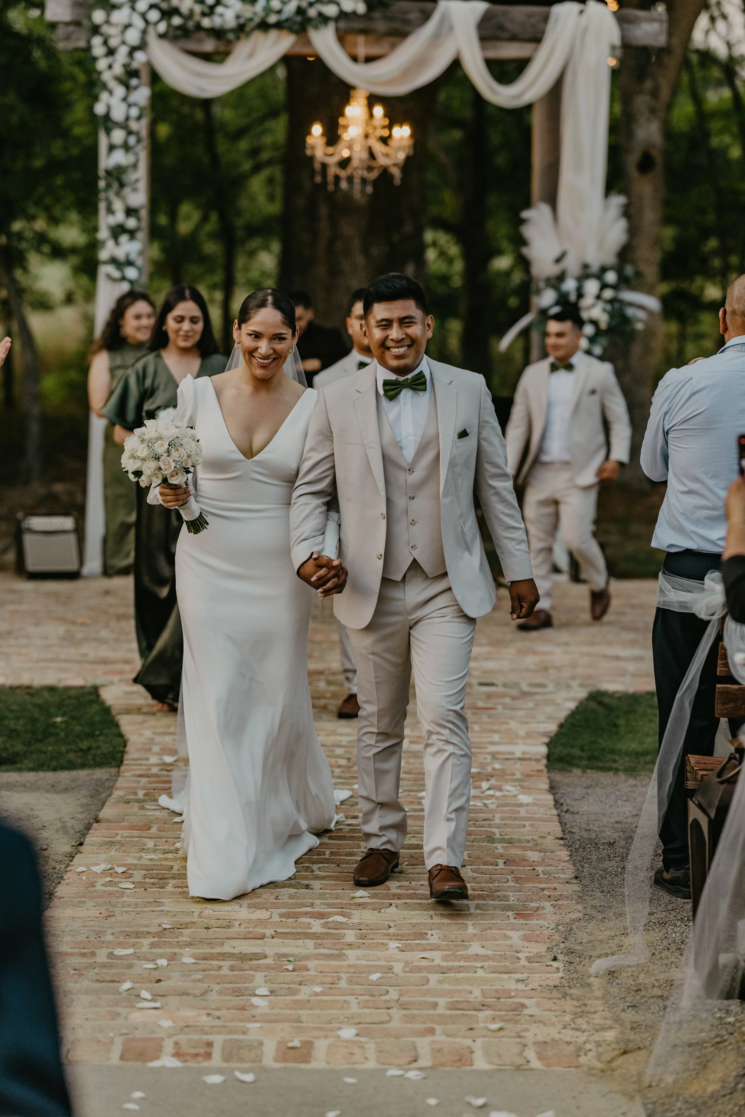 A newlywed couple walks down the aisle holding hands, smiling. The bride is in a white wedding dress holding a bouquet, and the groom is in a light-colored suit with a green bow tie. Guests and wedding party are visible behind them, and the scene is 