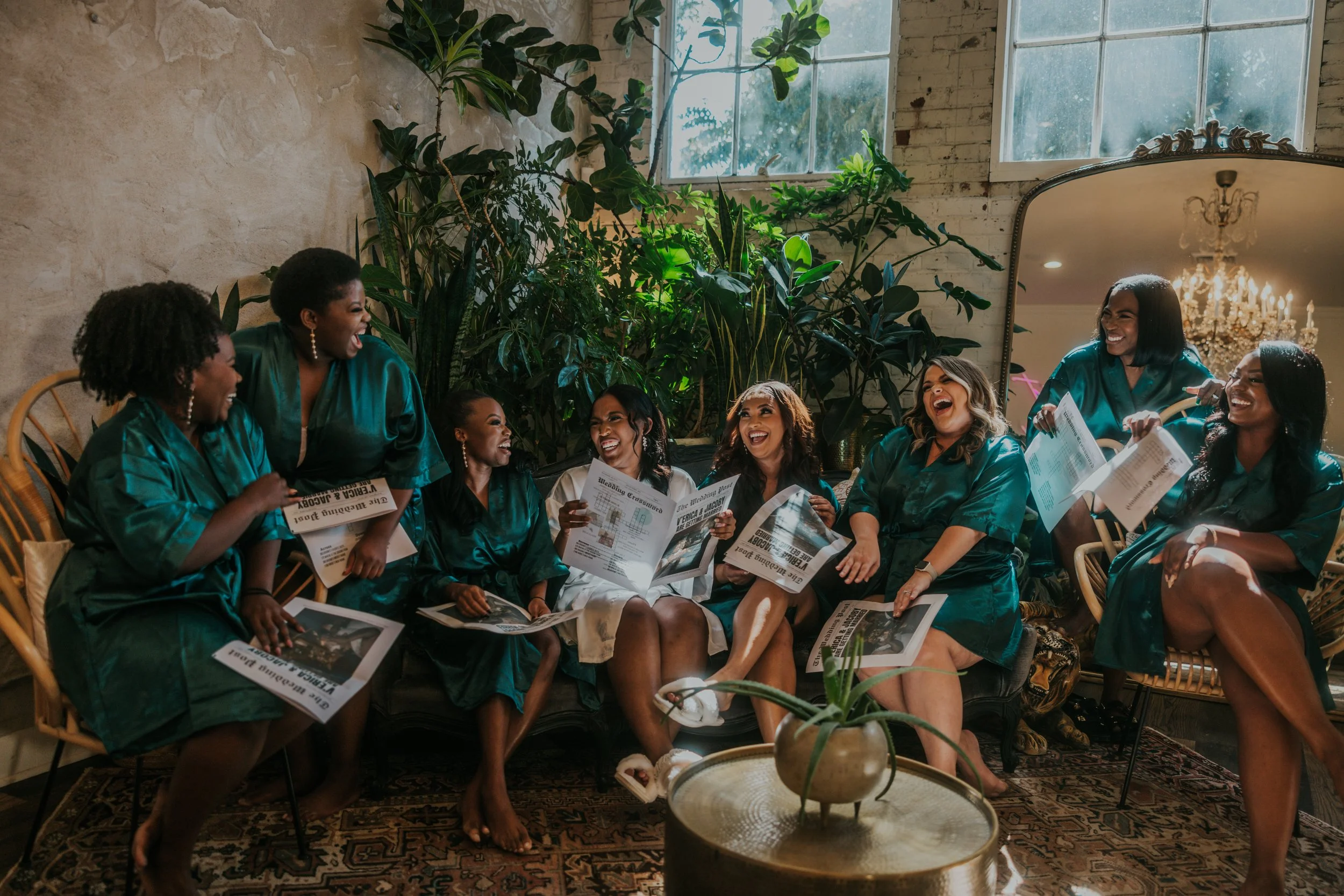 Group of women in matching satin robes laughing and chatting in a cozy, plant-filled room with a large mirror and chandelier.