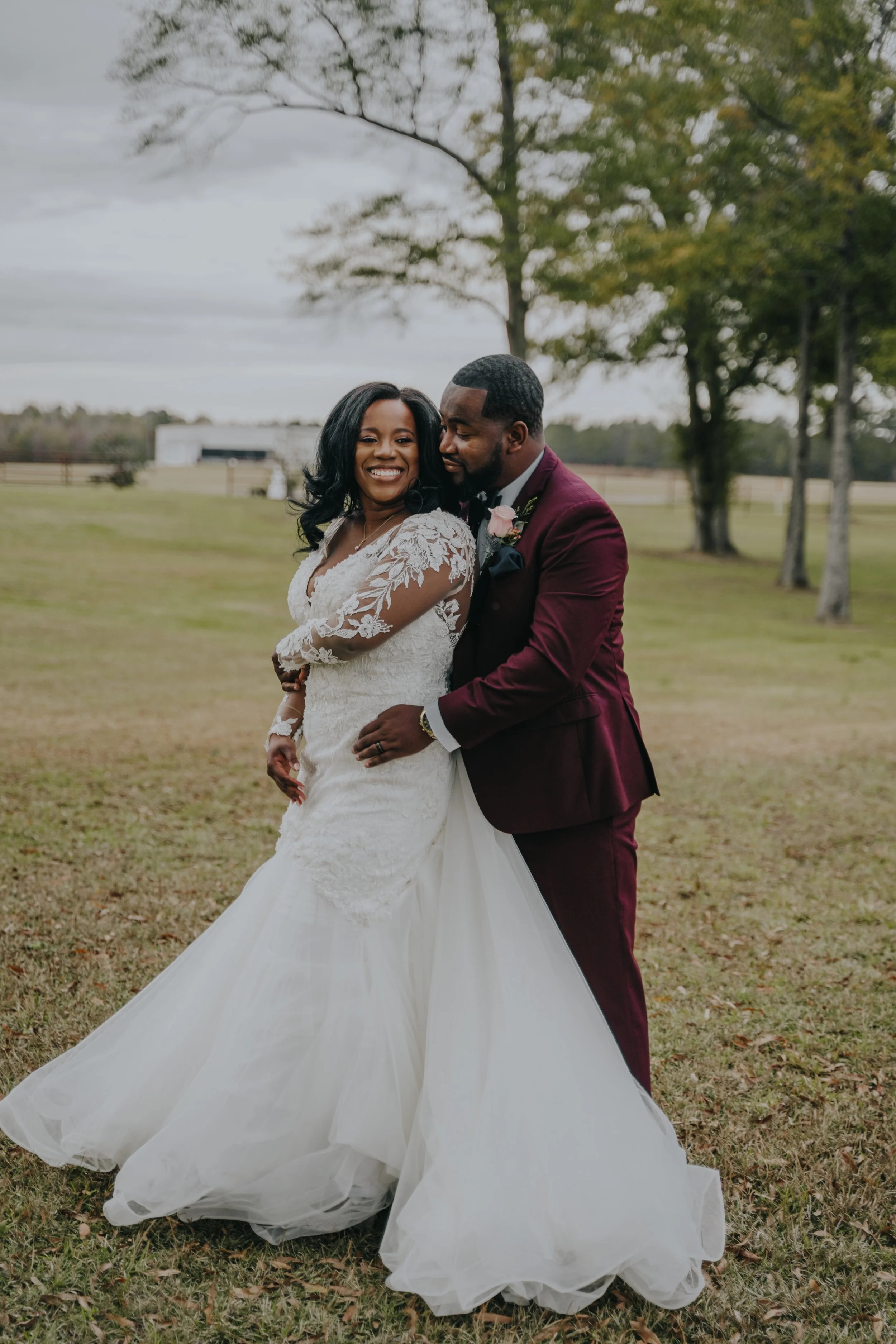 A smiling bride in a white wedding dress with lace sleeves and a groom in a burgundy suit embrace outdoors during the daytime, with trees and a cloudy sky in the background.