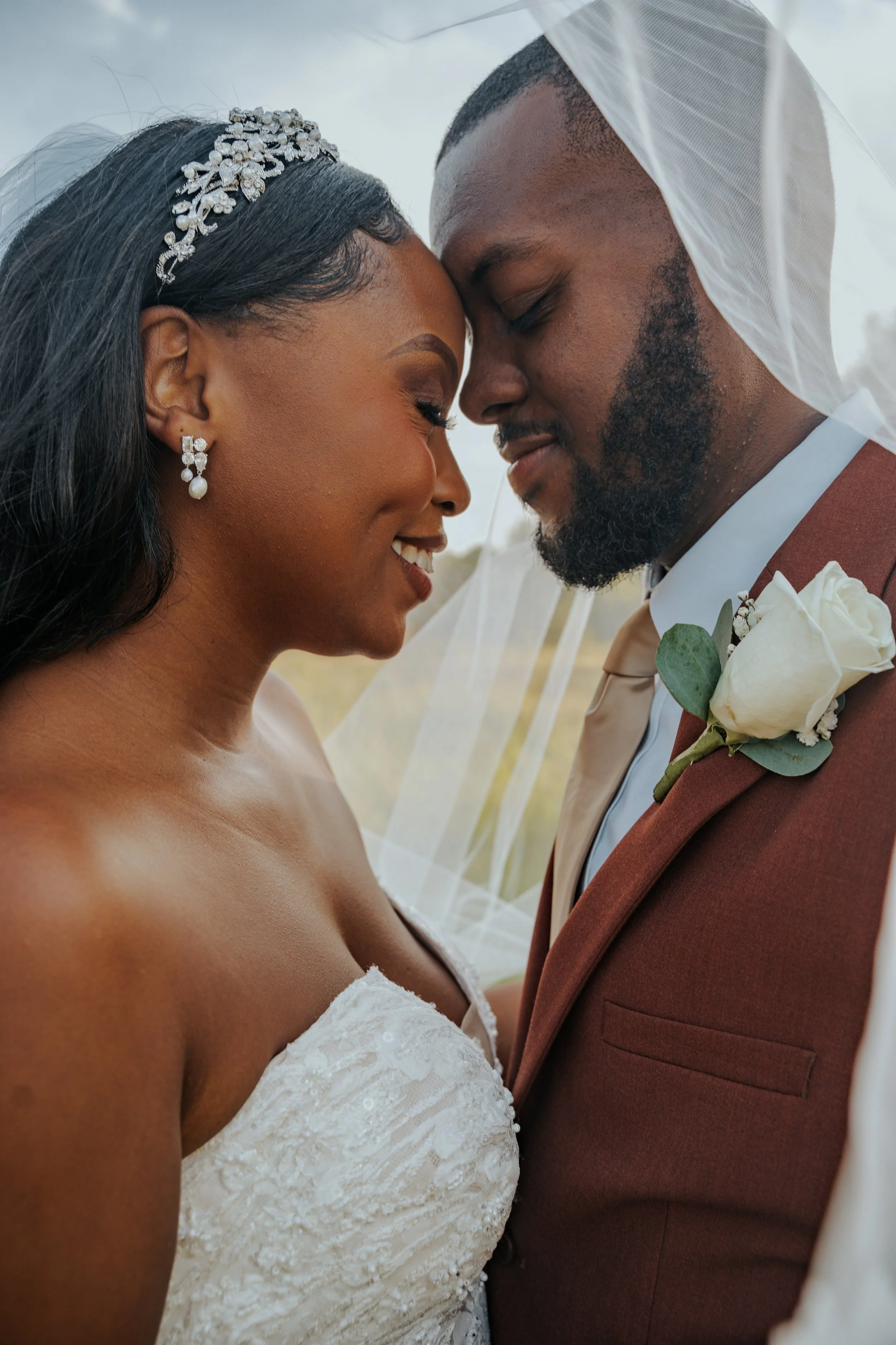 Close-up of a bride and groom with foreheads touching, smiling, outdoors with wedding attire, veil, and floral boutonniere.