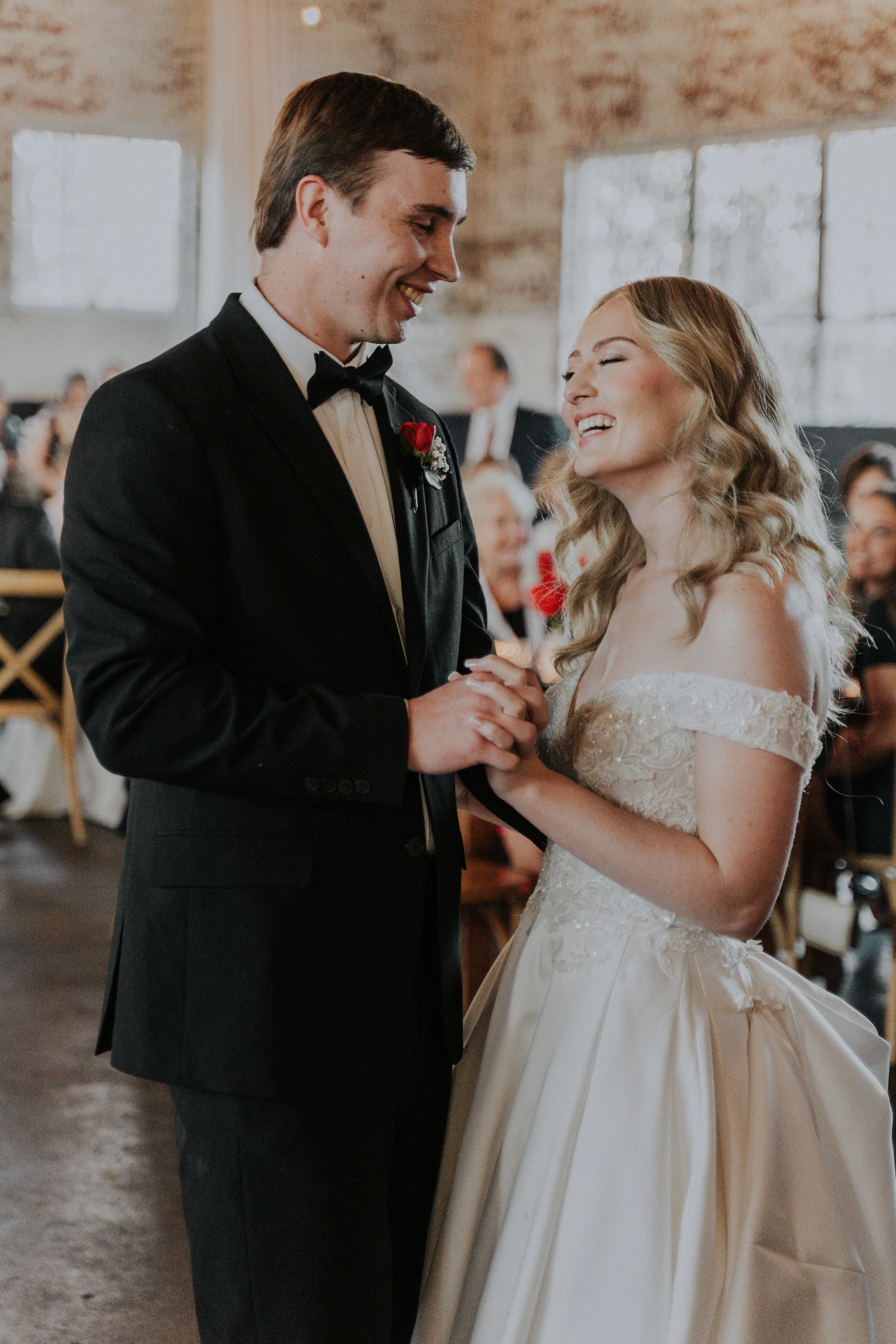 A bride and groom exchanging vows during their wedding ceremony in a rustic venue, holding hands and smiling.
