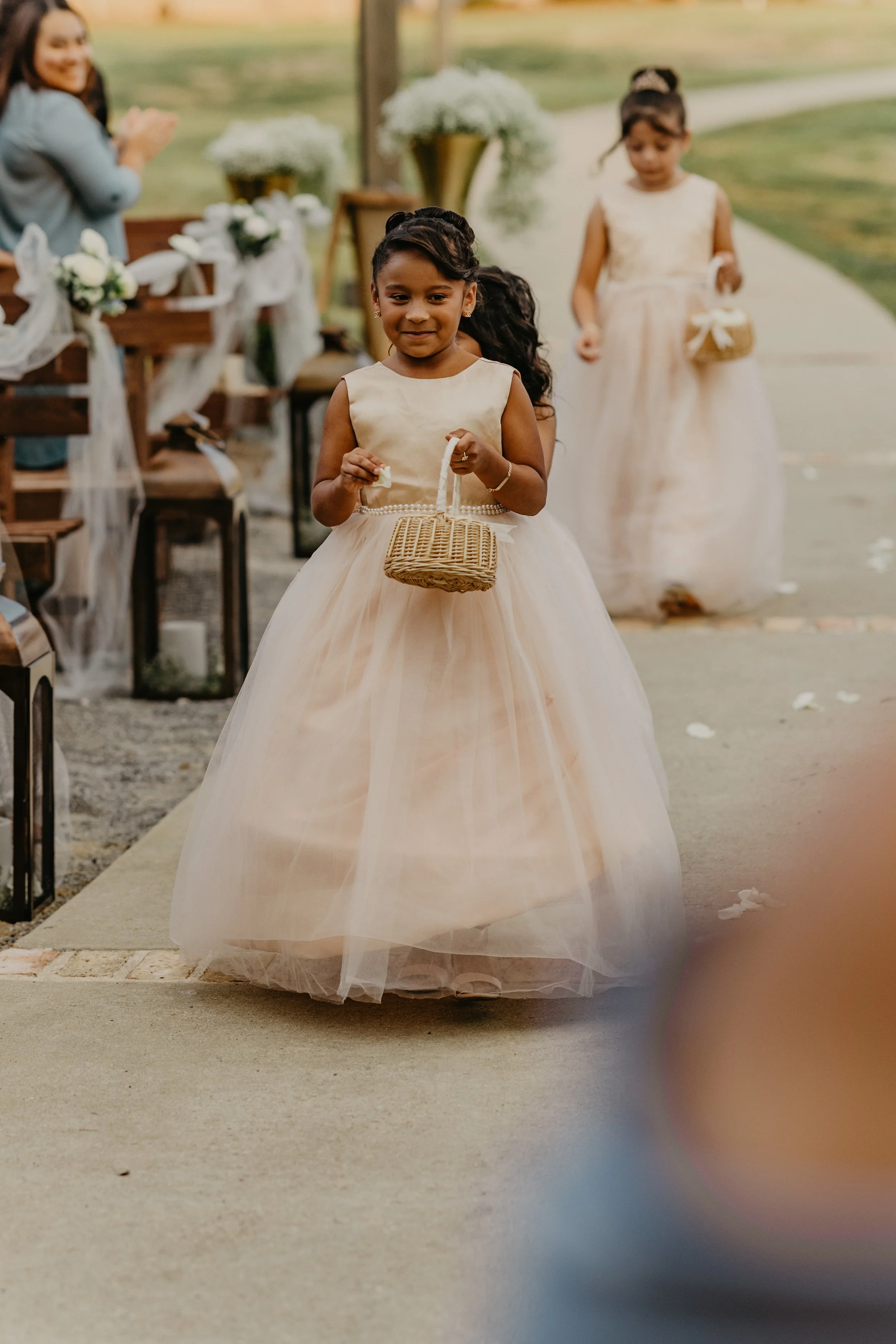 Young girl in a beige dress holding a small wicker basket walking down an outdoor aisle at a wedding ceremony, with other girls and guests in the background.