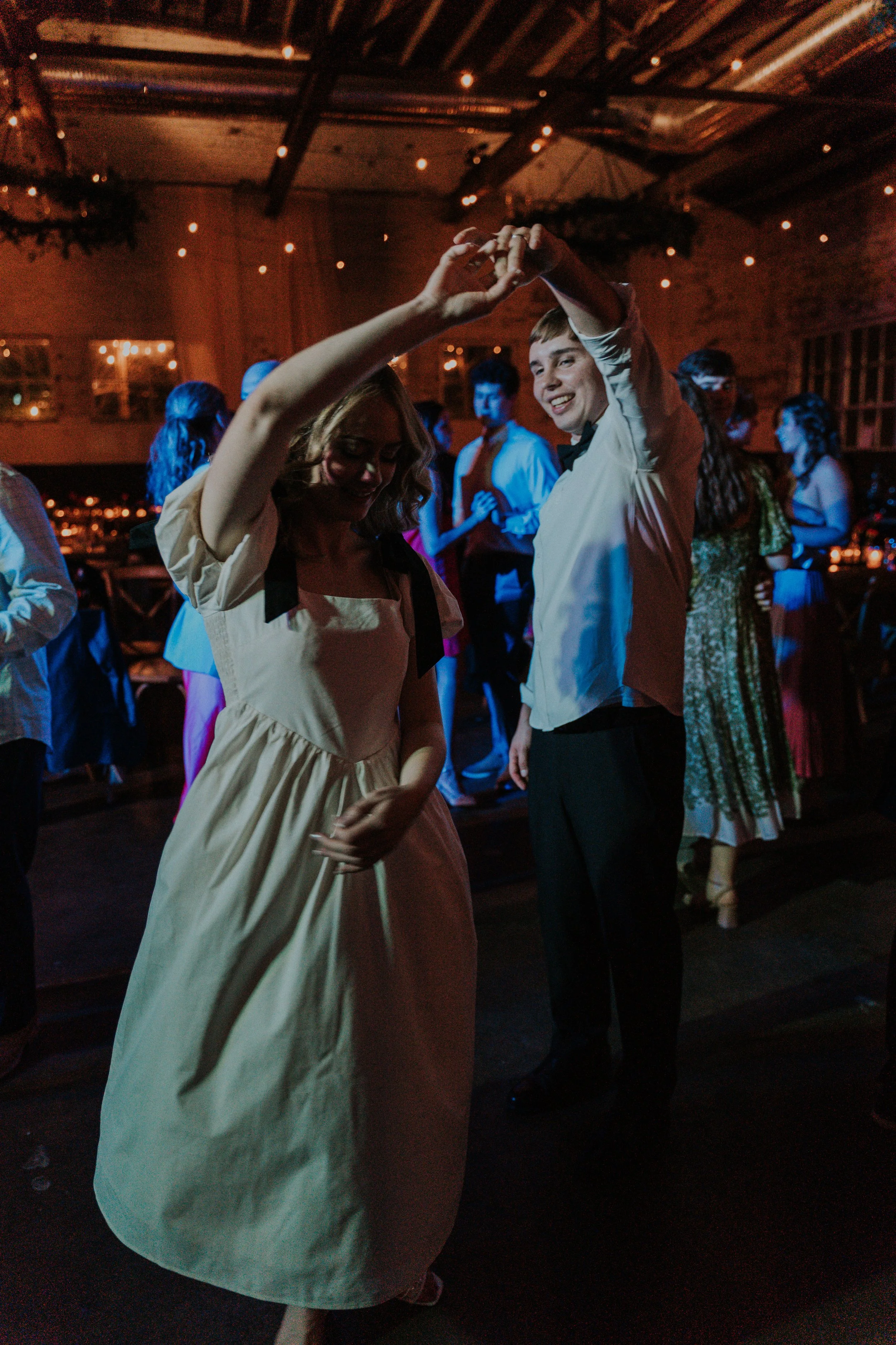 People dancing at a party or wedding reception, with a couple in the foreground rotating hands and smiling, under warm lighting in a rustic venue.