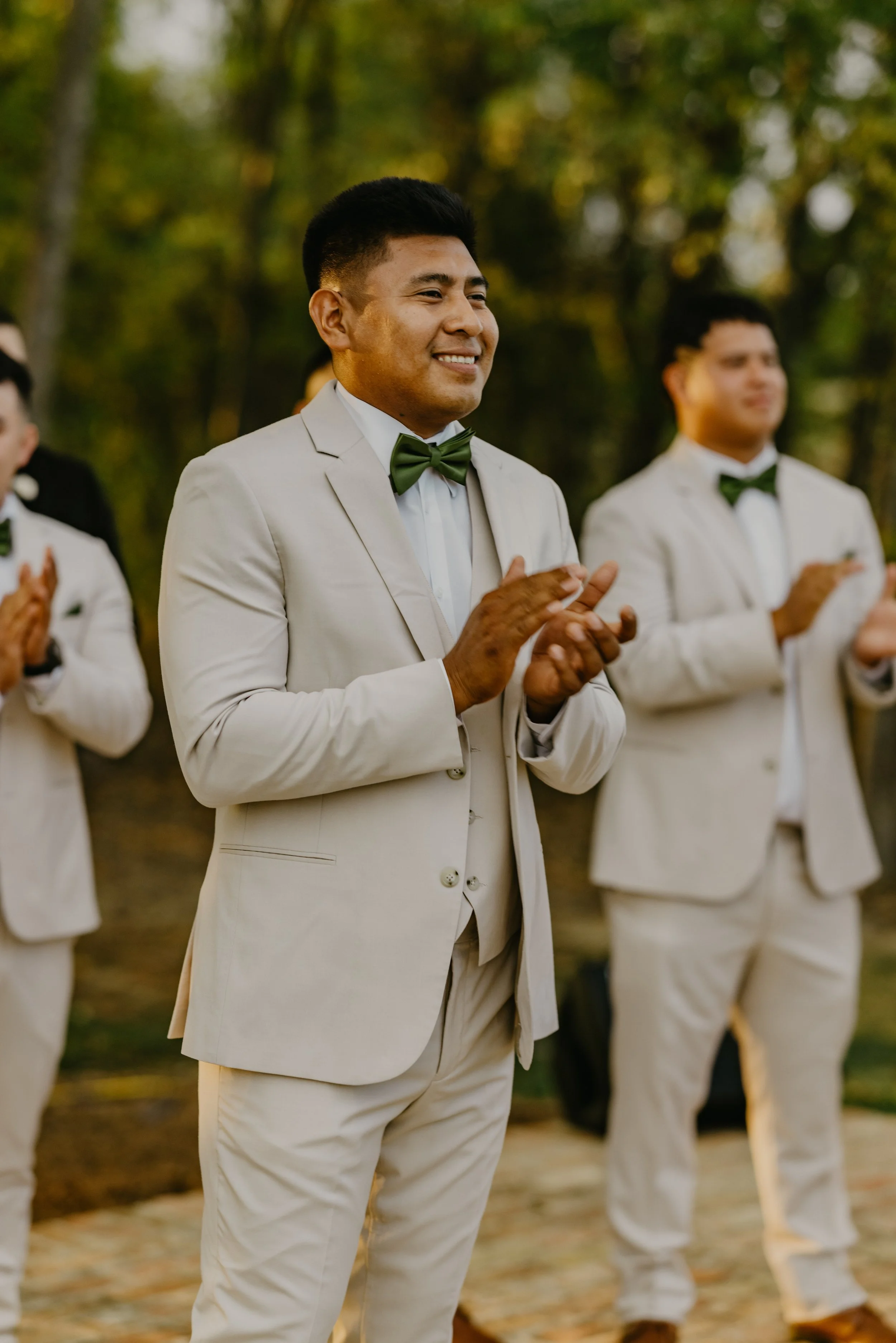 Group of men in cream suits and green bow ties standing outdoors, clapping at a wedding ceremony.