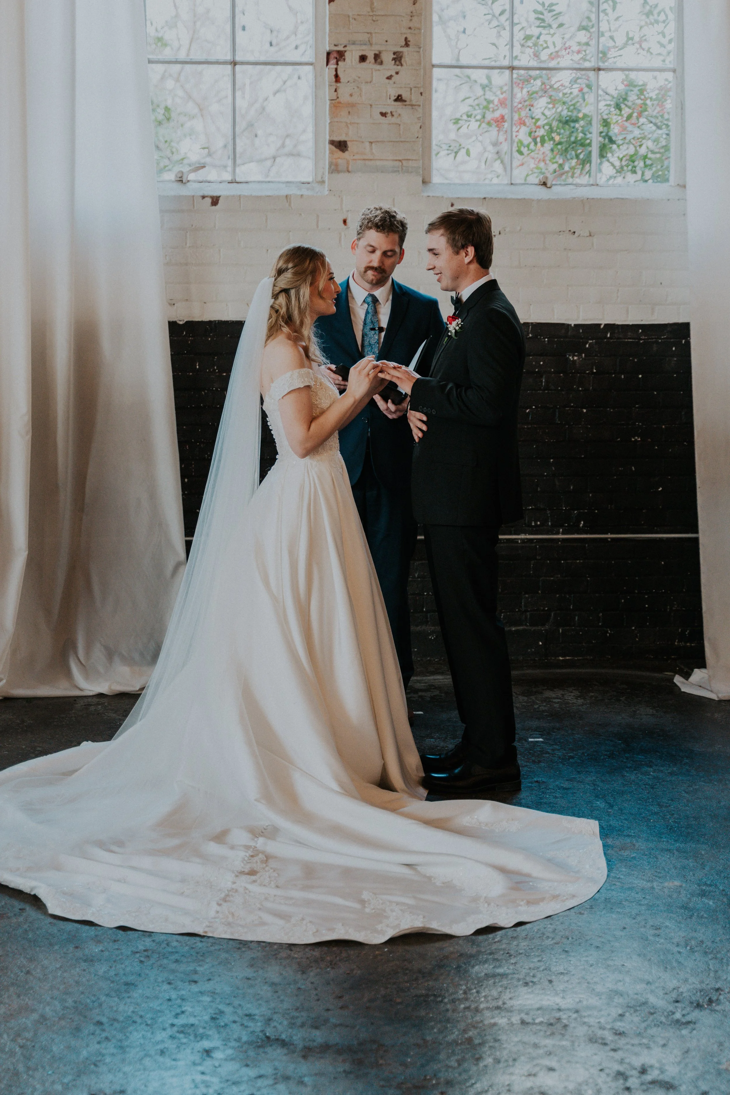A bride and groom exchanging vows during a wedding ceremony in an industrial-style venue with exposed brick, large windows, and natural light.