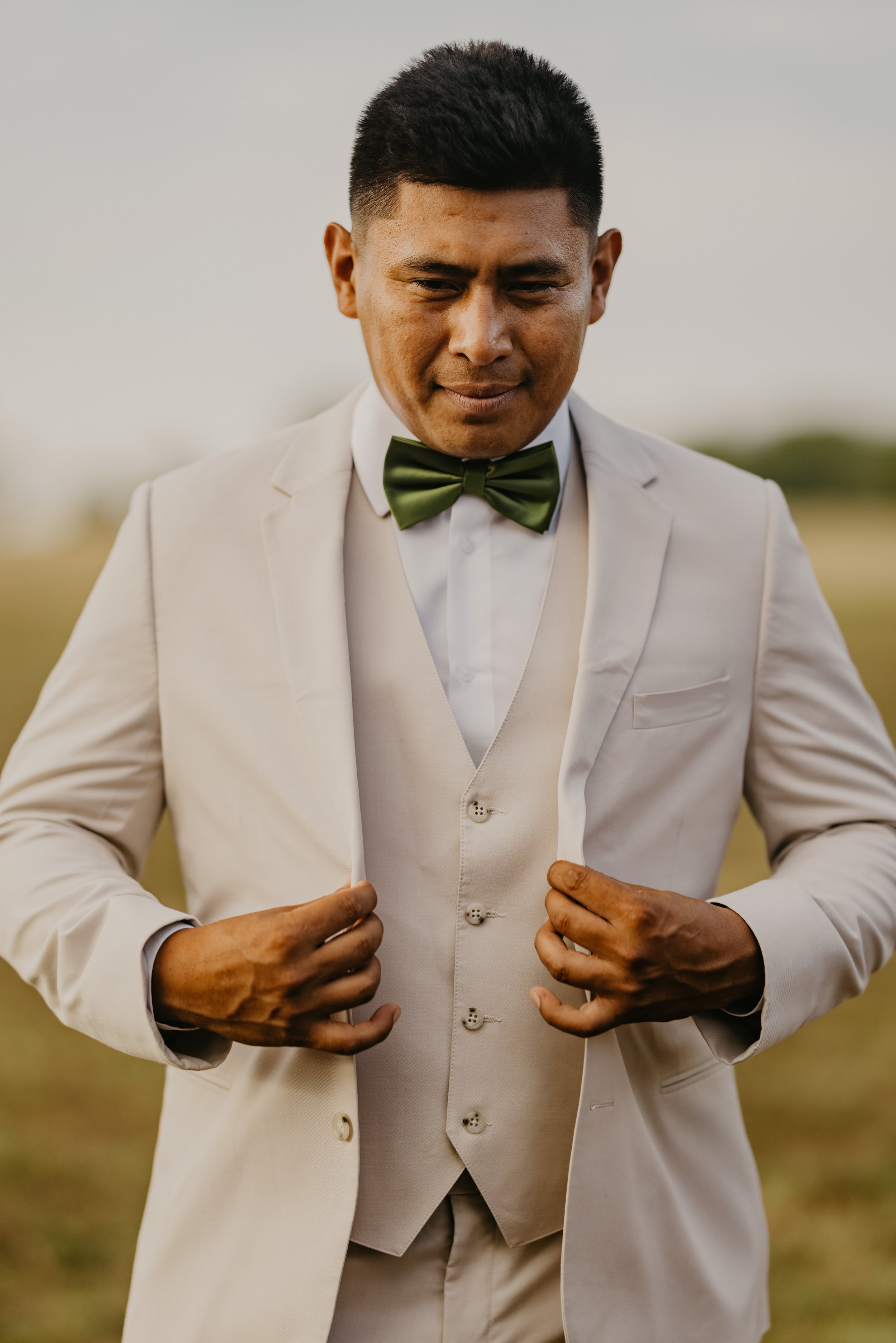 A man in a white tuxedo with a green bow tie adjusting his jacket outdoors.