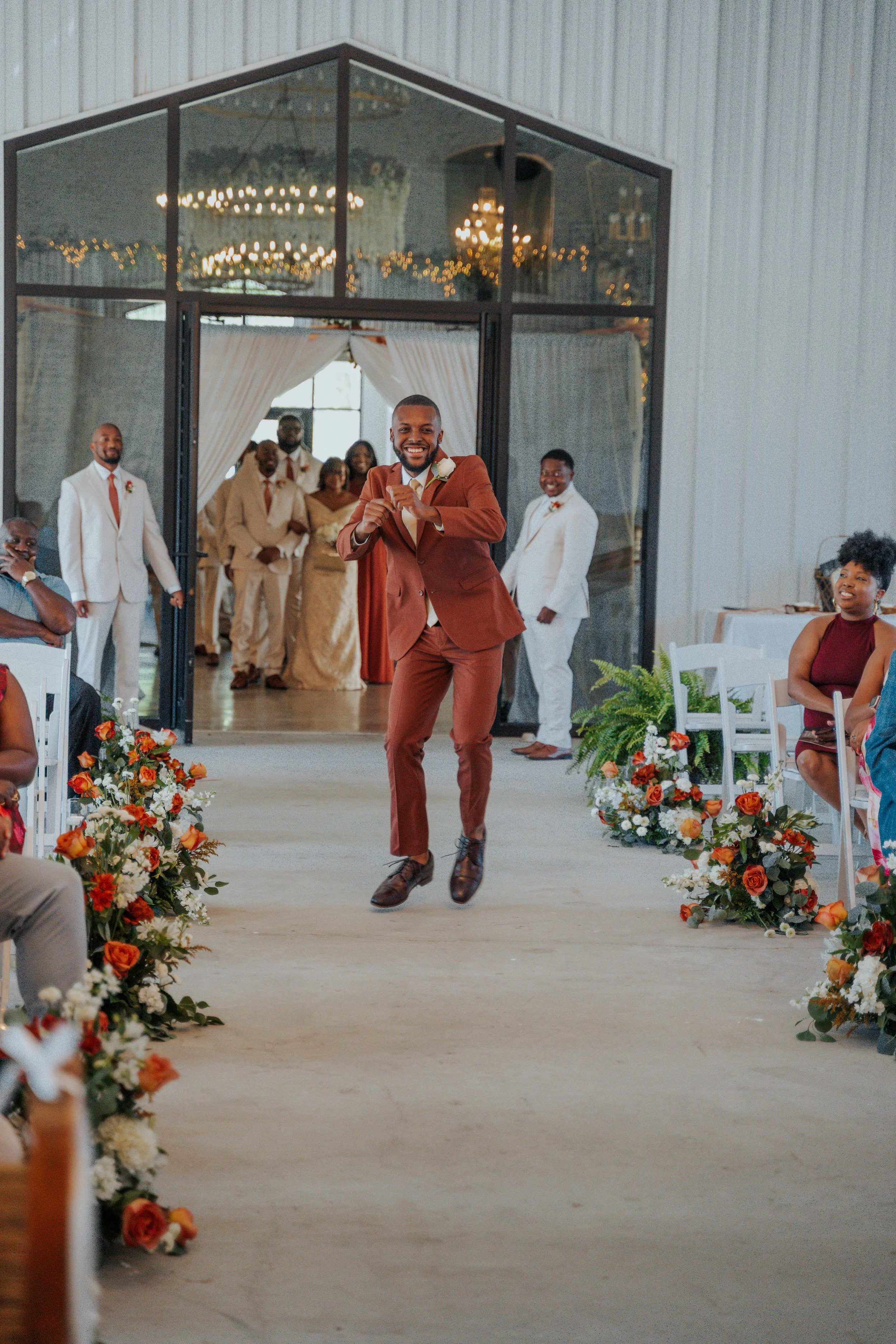 A man in a rust-colored suit walks down an aisle at a wedding celebration, smiling and adjusting his watch. Guests seated on either side are dressed in formal attire, with floral arrangements lining the aisle. In the background, the wedding party sta
