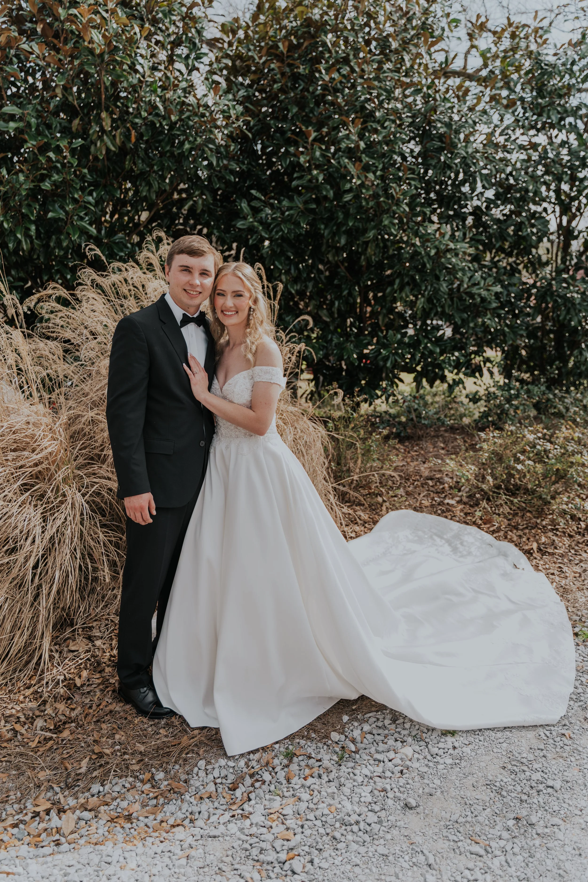 A newlywed couple standing outdoors, the groom in a black tuxedo and the bride in a white wedding gown, smiling together.