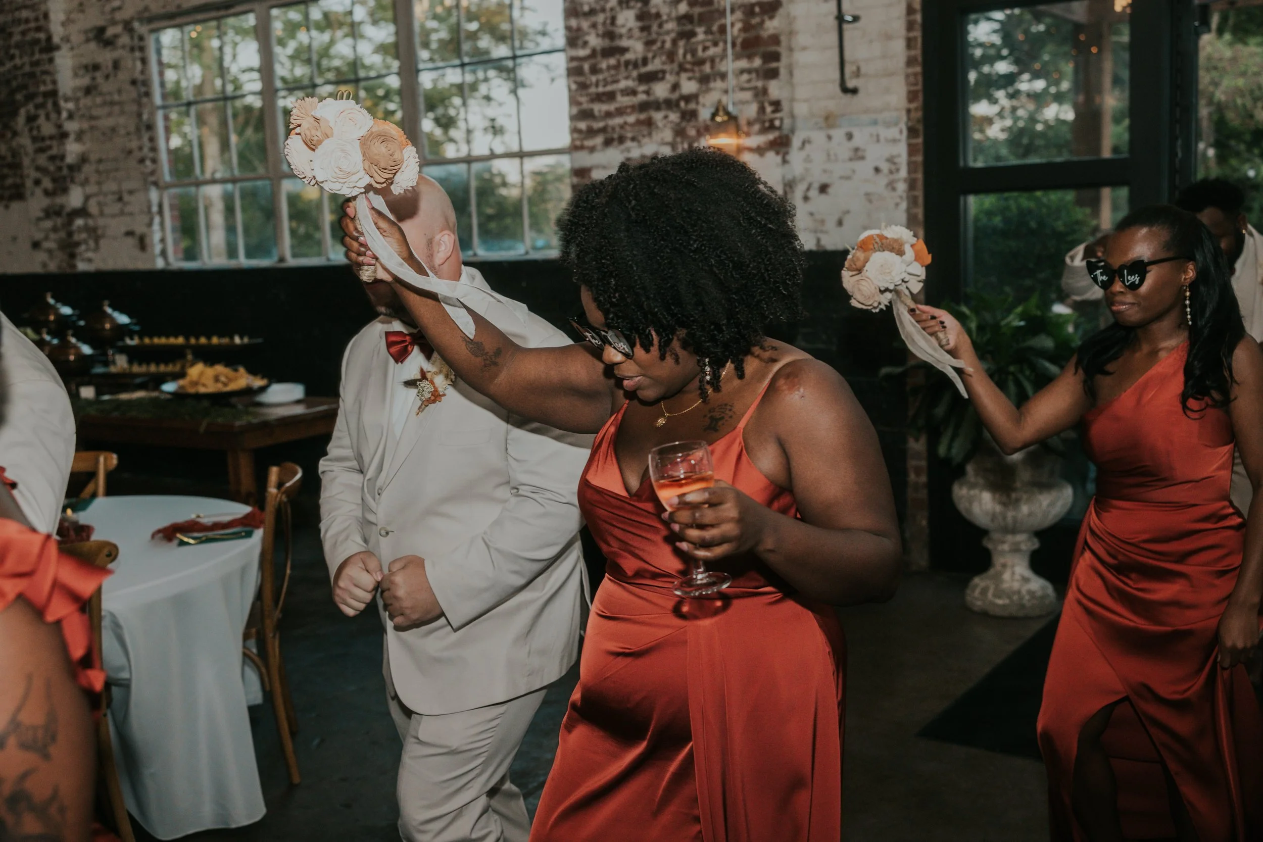 People dancing at a wedding reception, wearing formal attire including orange dresses and a white tuxedo with a red bow tie, with a rustic industrial-style interior and large windows in the background.