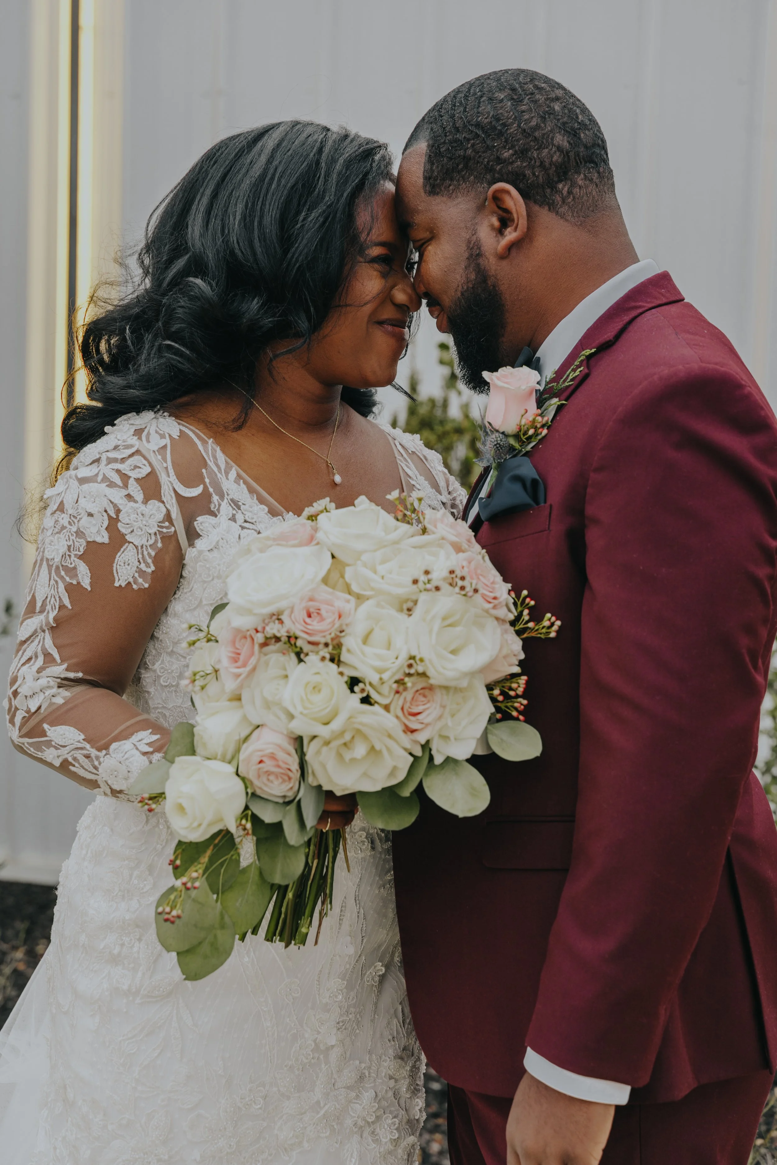 A bride and groom nose touching, holding a bouquet of white and pink roses, in a wedding setting.