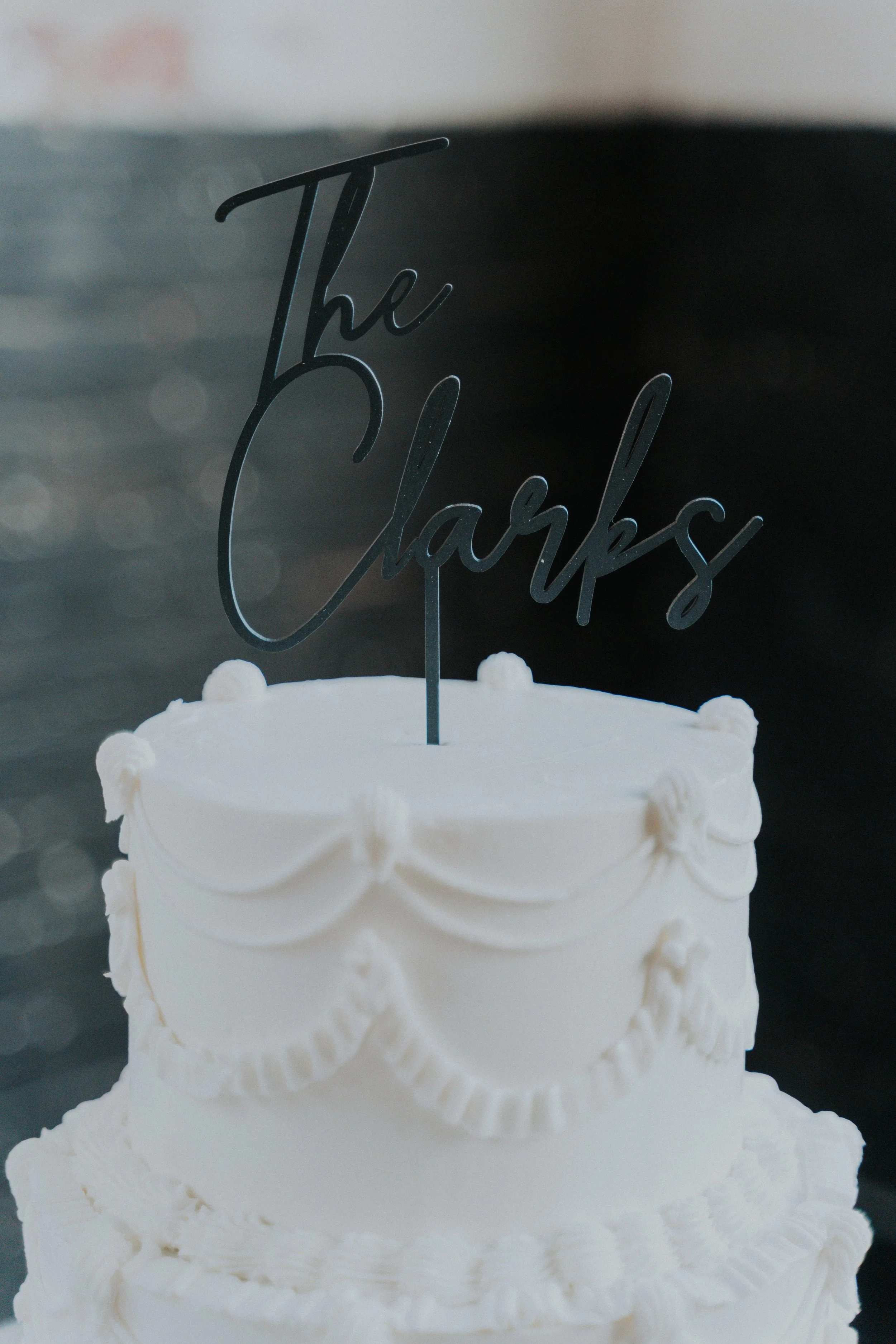 A white cake with decorative piping and a black cake topper that says 'The Clarks' on top.