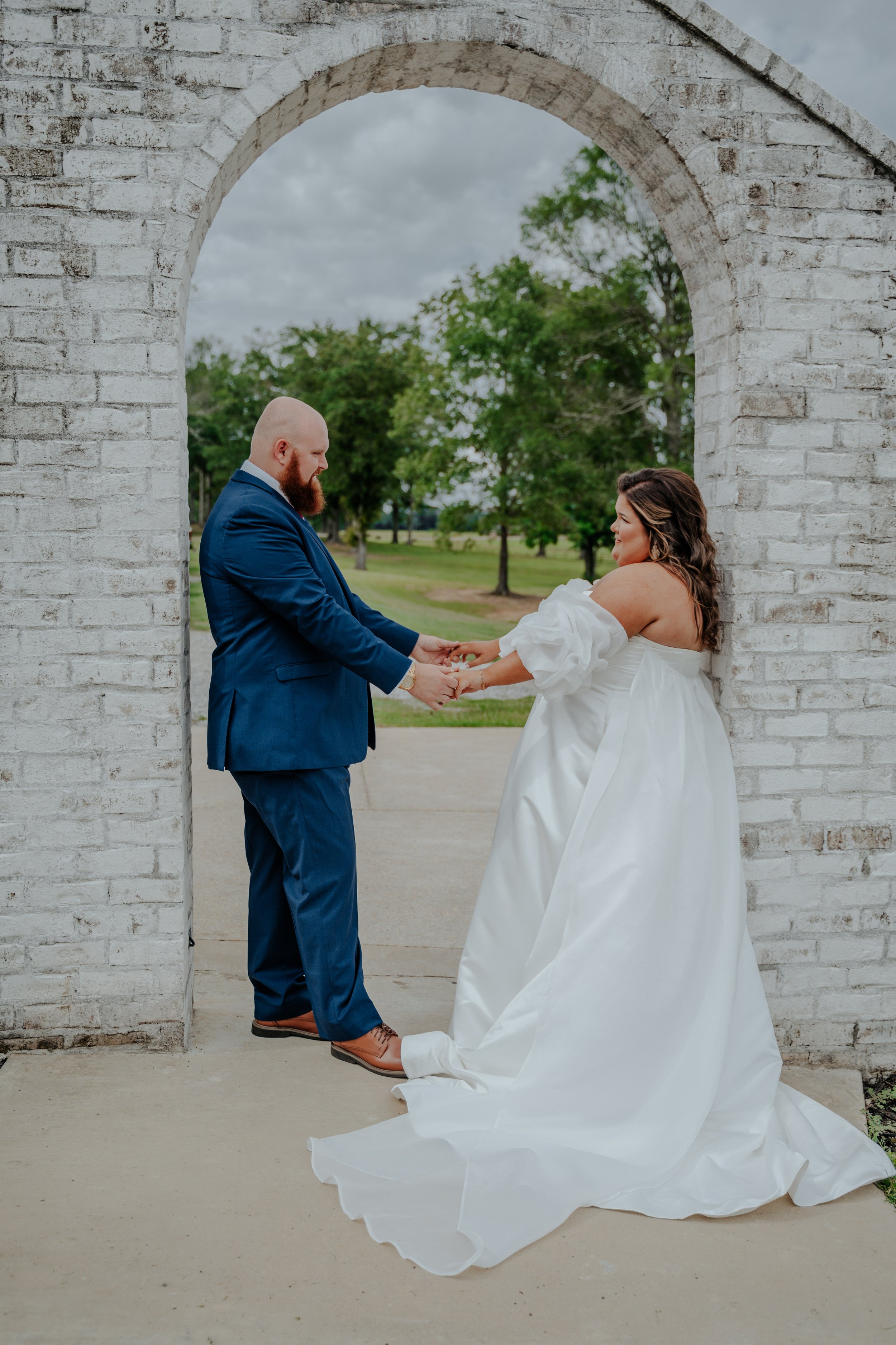 A bride and groom holding hands during their outdoor wedding ceremony, standing under a white brick archway with green trees and cloudy sky in the background.
