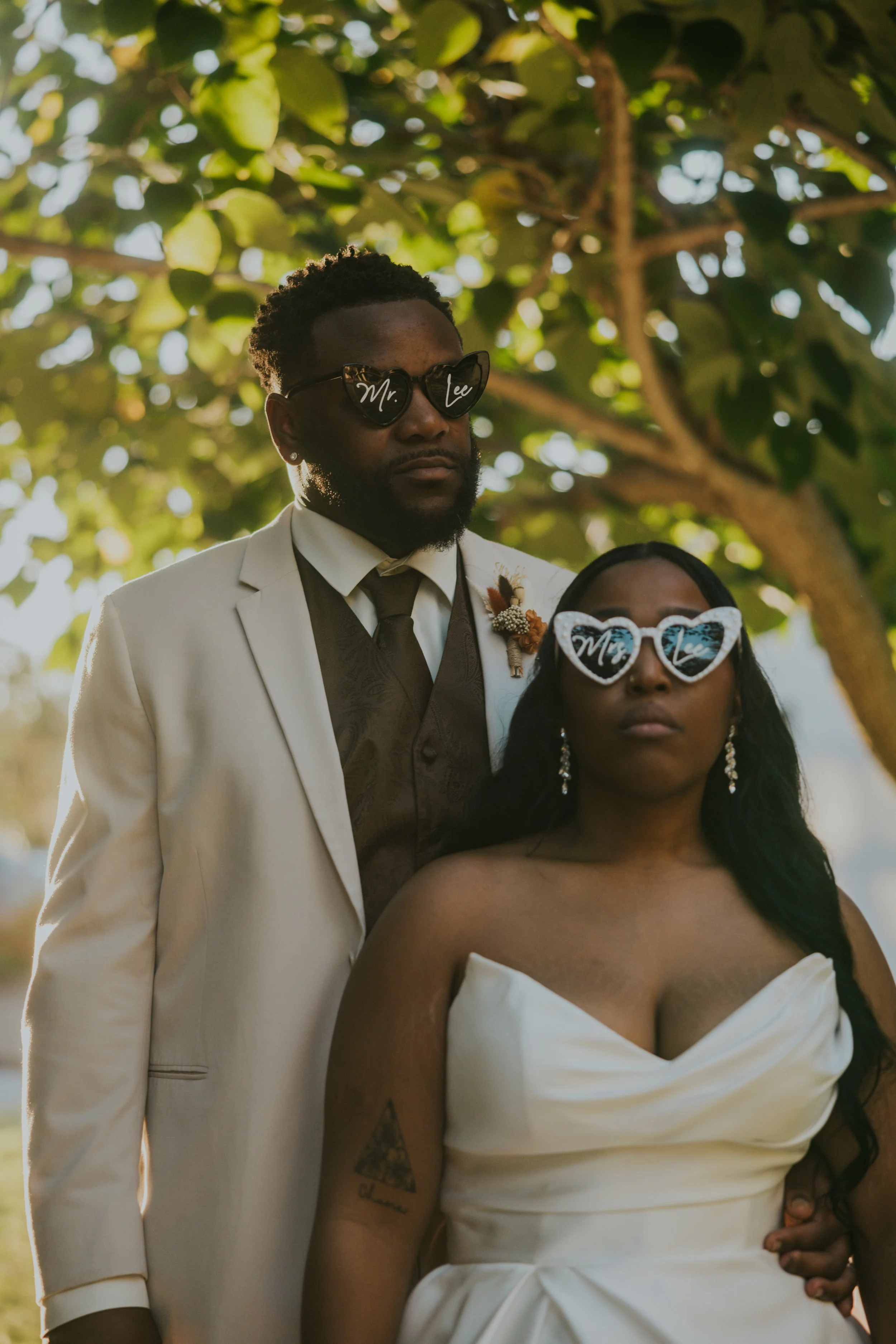 A couple dressed in wedding attire standing outdoors under a tree with green leaves, wearing matching sunglasses with 'Mr' and 'Mrs' written on them.