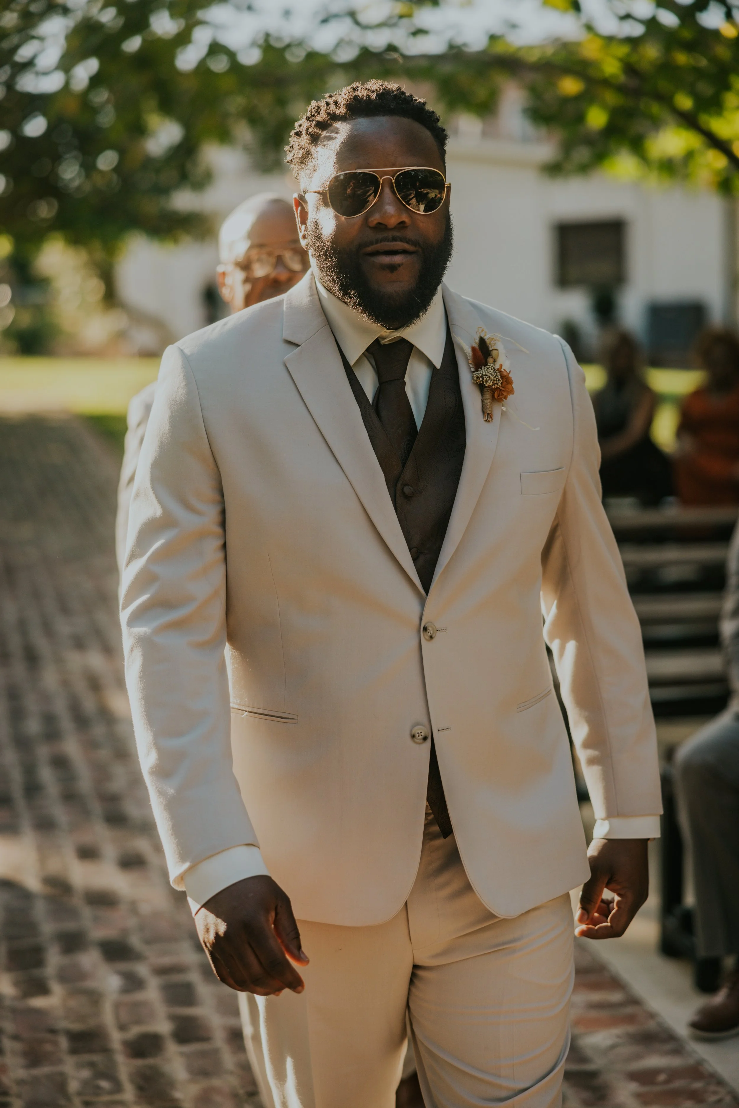 Man in a beige suit and sunglasses standing outdoors during a formal event, with other people and trees in the background.