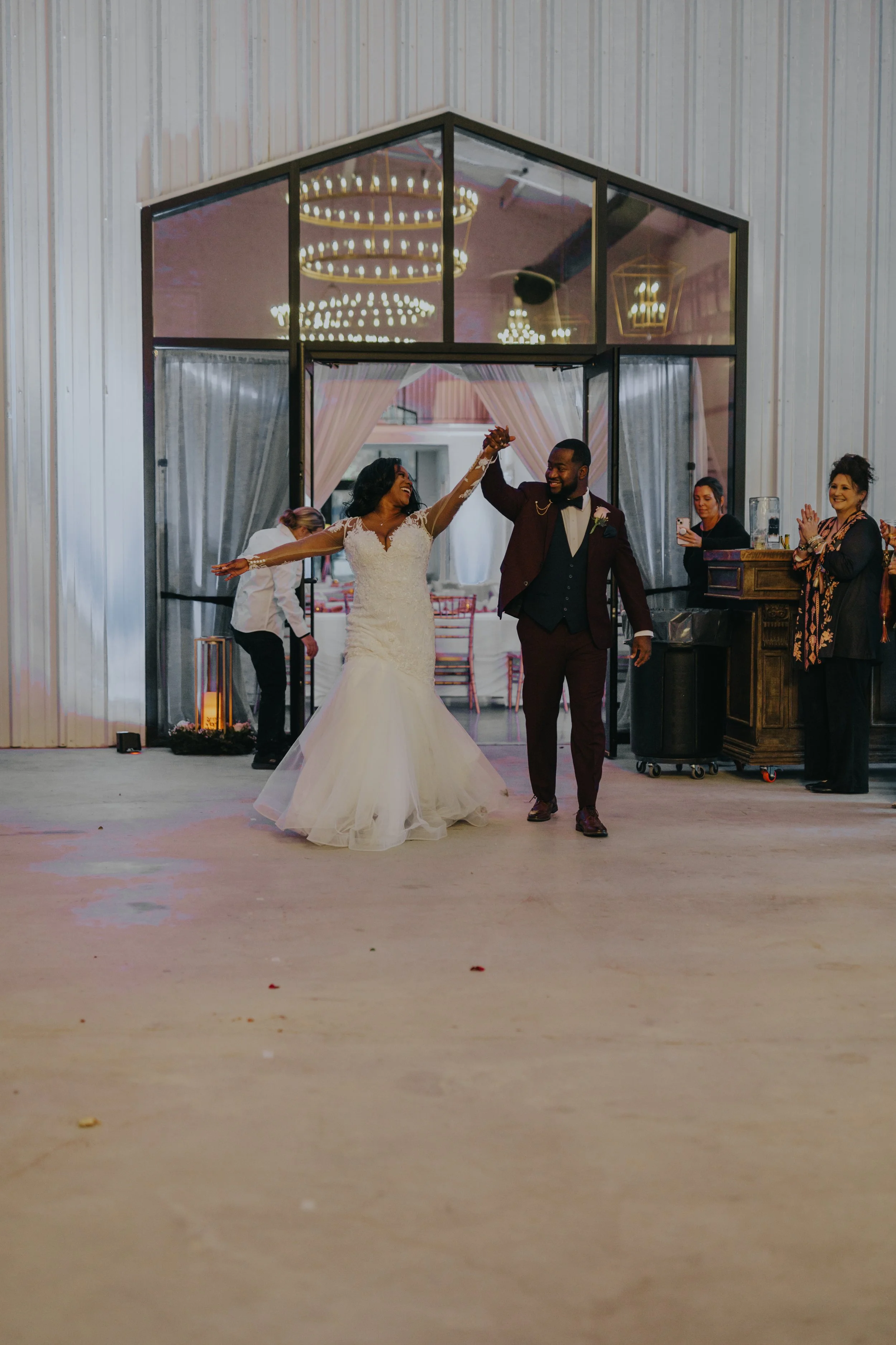 A bride and groom dancing at their wedding reception indoors, surrounded by guests, with chandeliers hanging overhead.