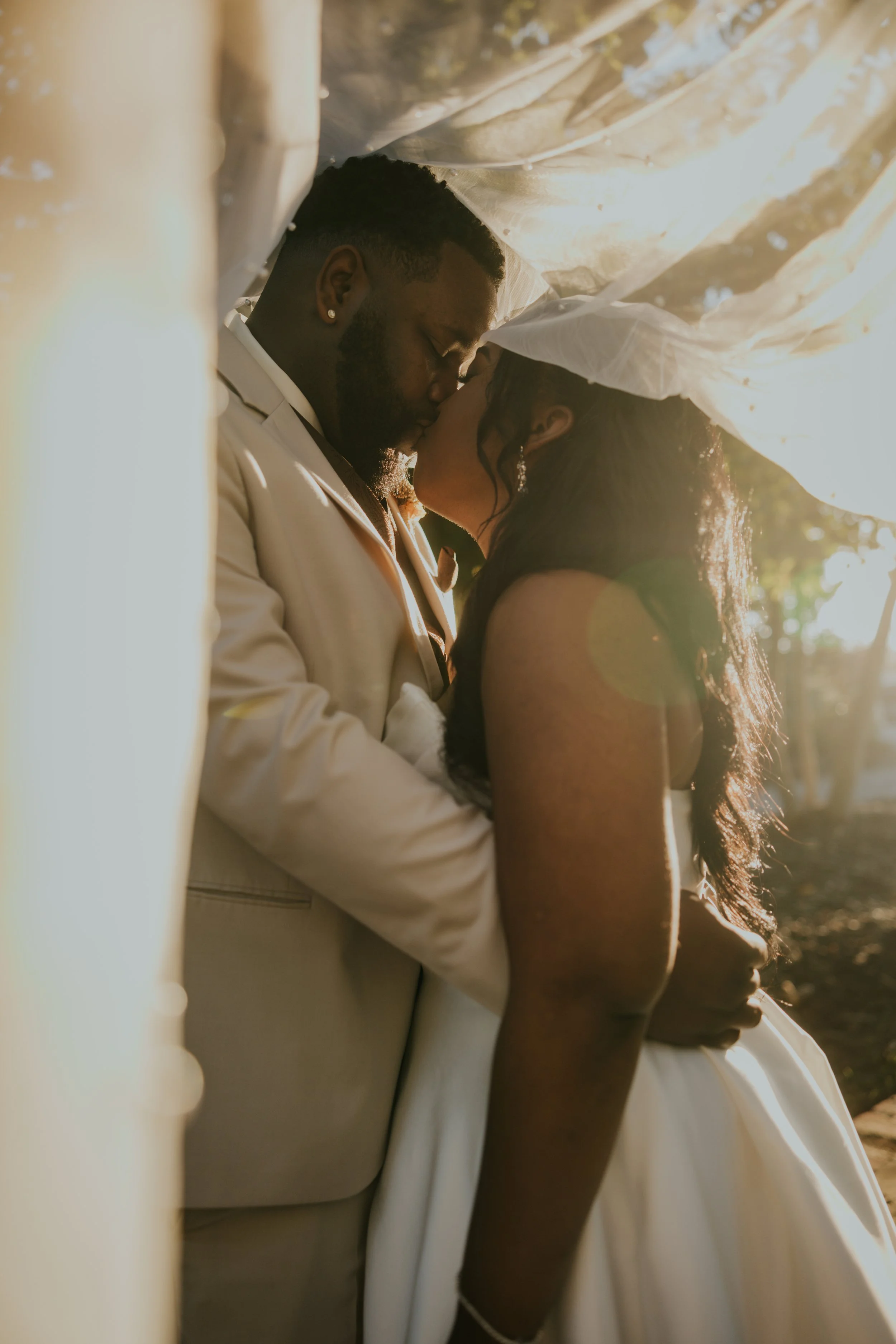 A couple kissing under a canopy, wearing wedding attire, with sunlight filtering through the fabric.