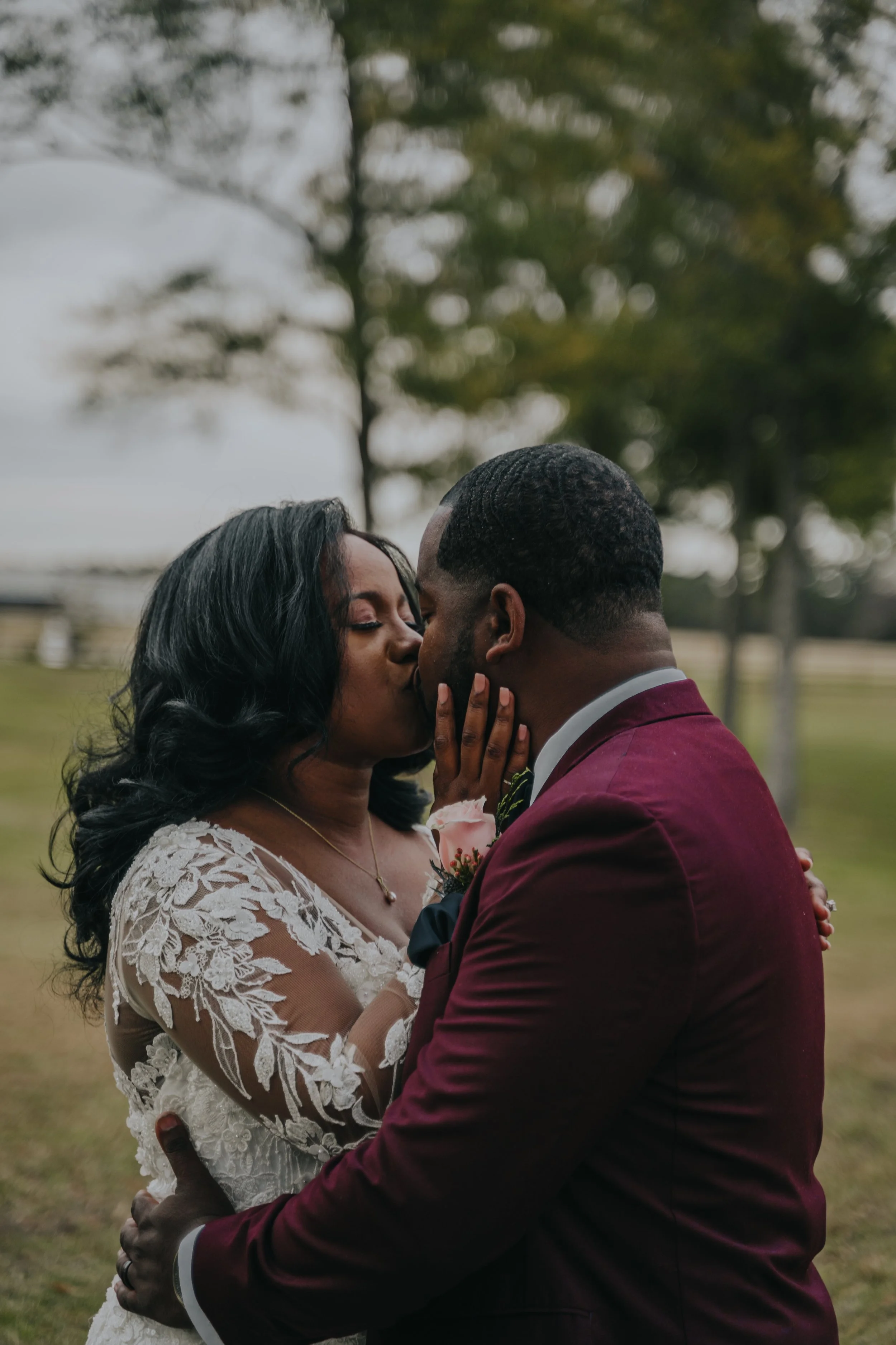 A couple sharing a kiss outdoors, dressed in wedding attire, with trees and a cloudy sky in the background.