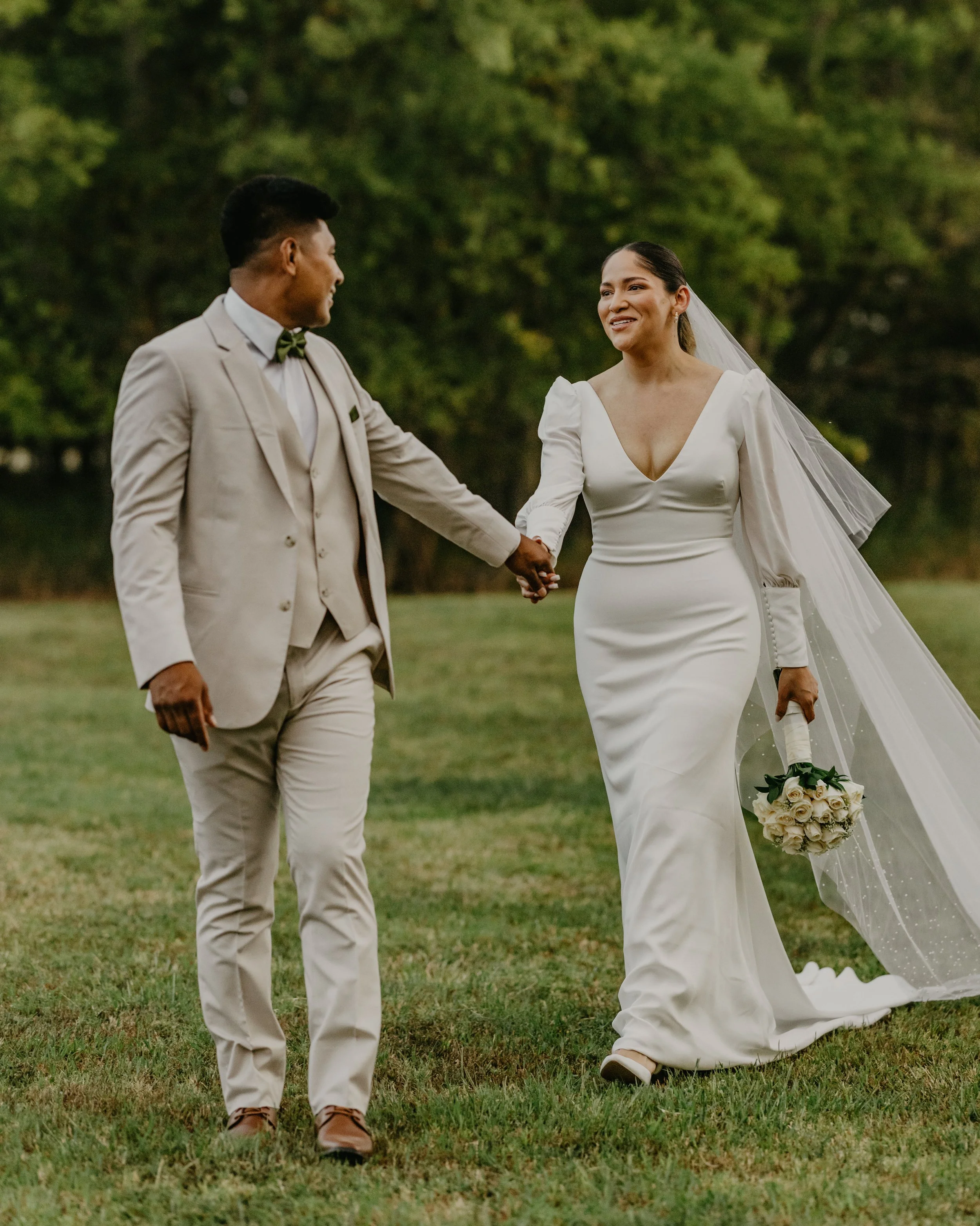 A couple dressed in wedding attire holding hands outdoors, the bride in a white gown with long sleeves and a veil, holding a bouquet of white roses, and the groom in a beige suit with a green bow tie, walking on grass with a background of trees.