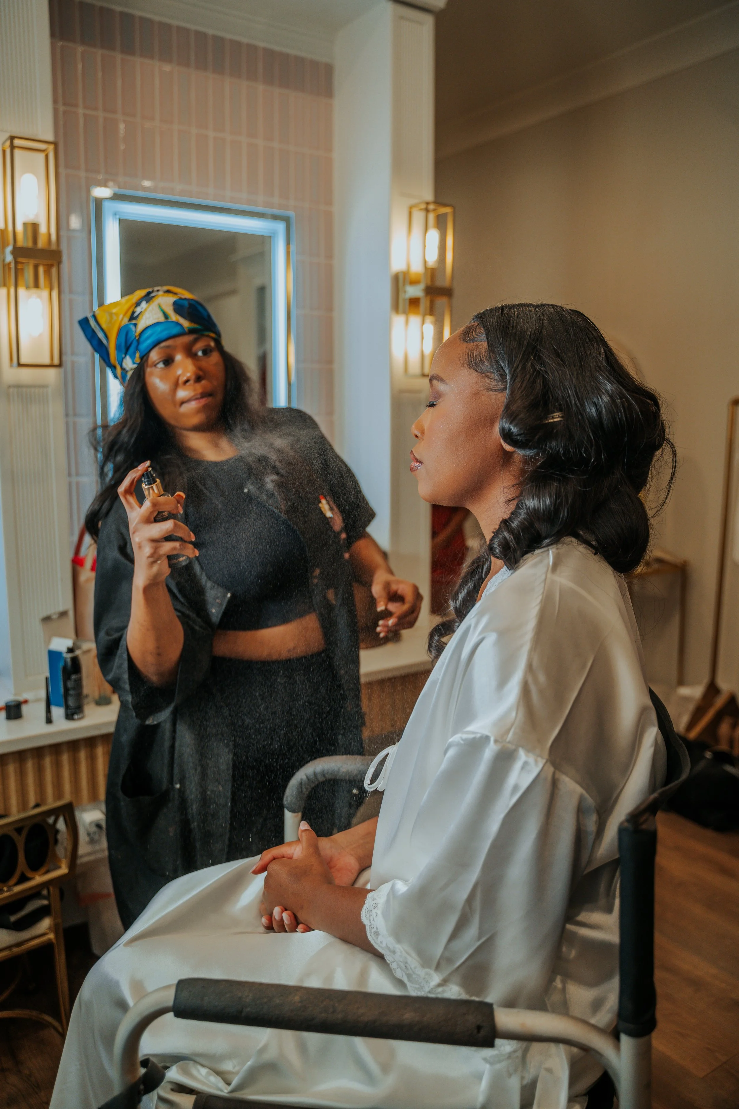 Woman in a wheelchair getting her hair styled while another woman sprays her with perfume in a well-lit room.