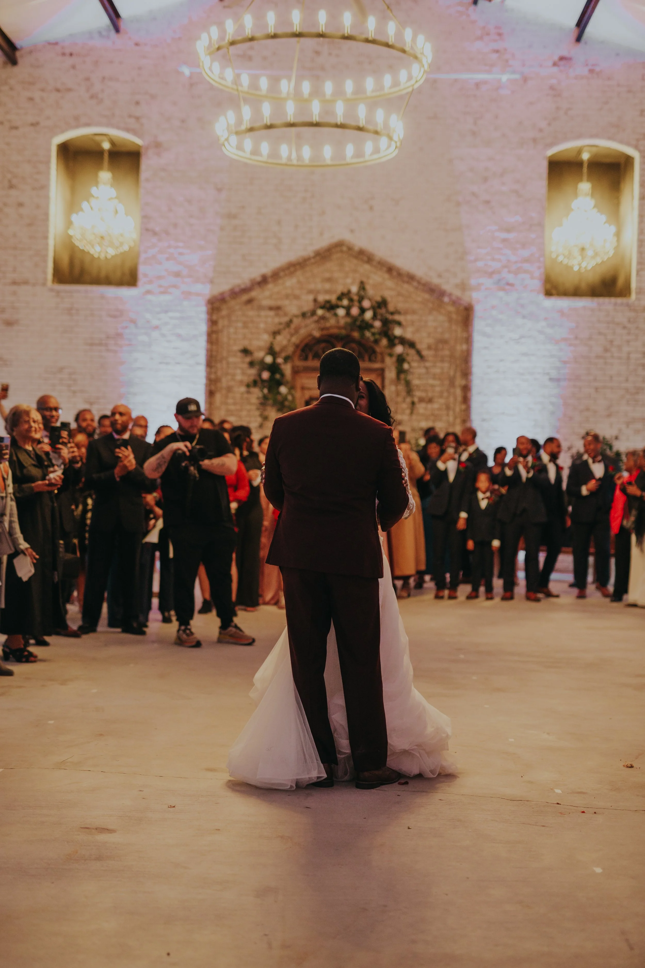 A bride and groom share their first dance at their wedding reception. The couple is facing each other, surrounded by guests in a rustic, well-lit venue with chandeliers and exposed brick walls.