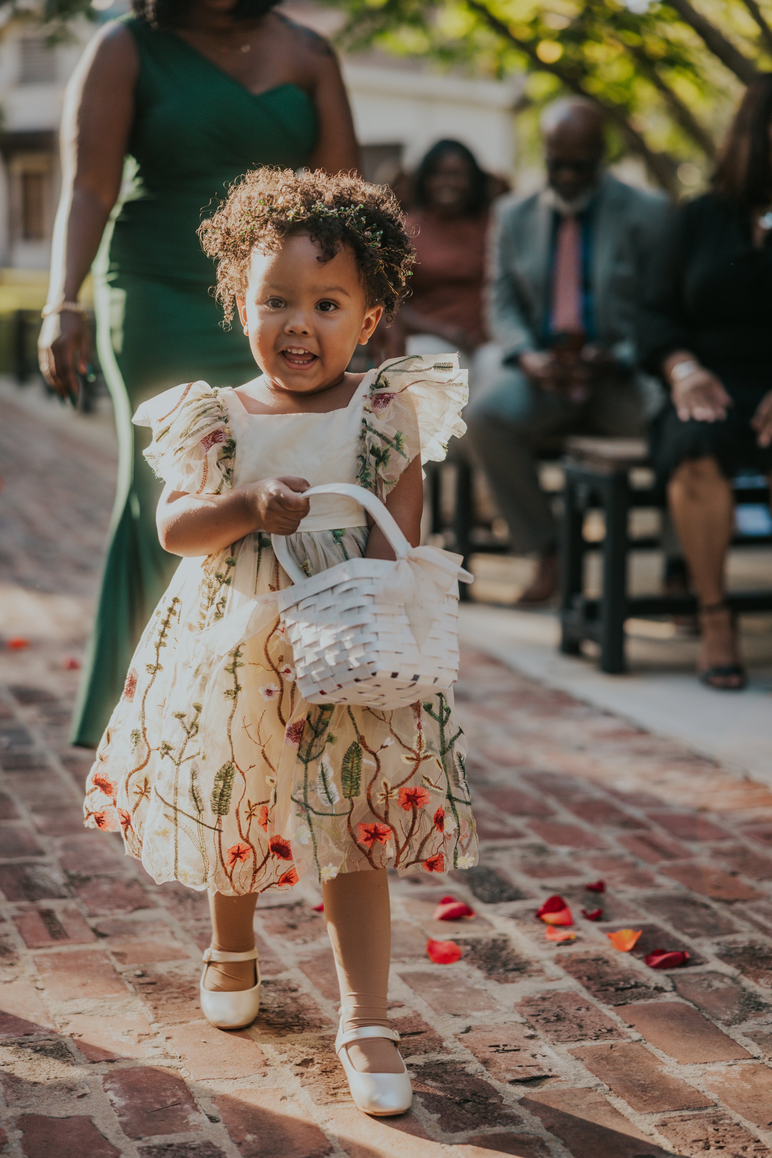 A young girl with curly hair in a floral dress carrying a small white basket, walking on a brick path decorated with flower petals during an outdoor event.