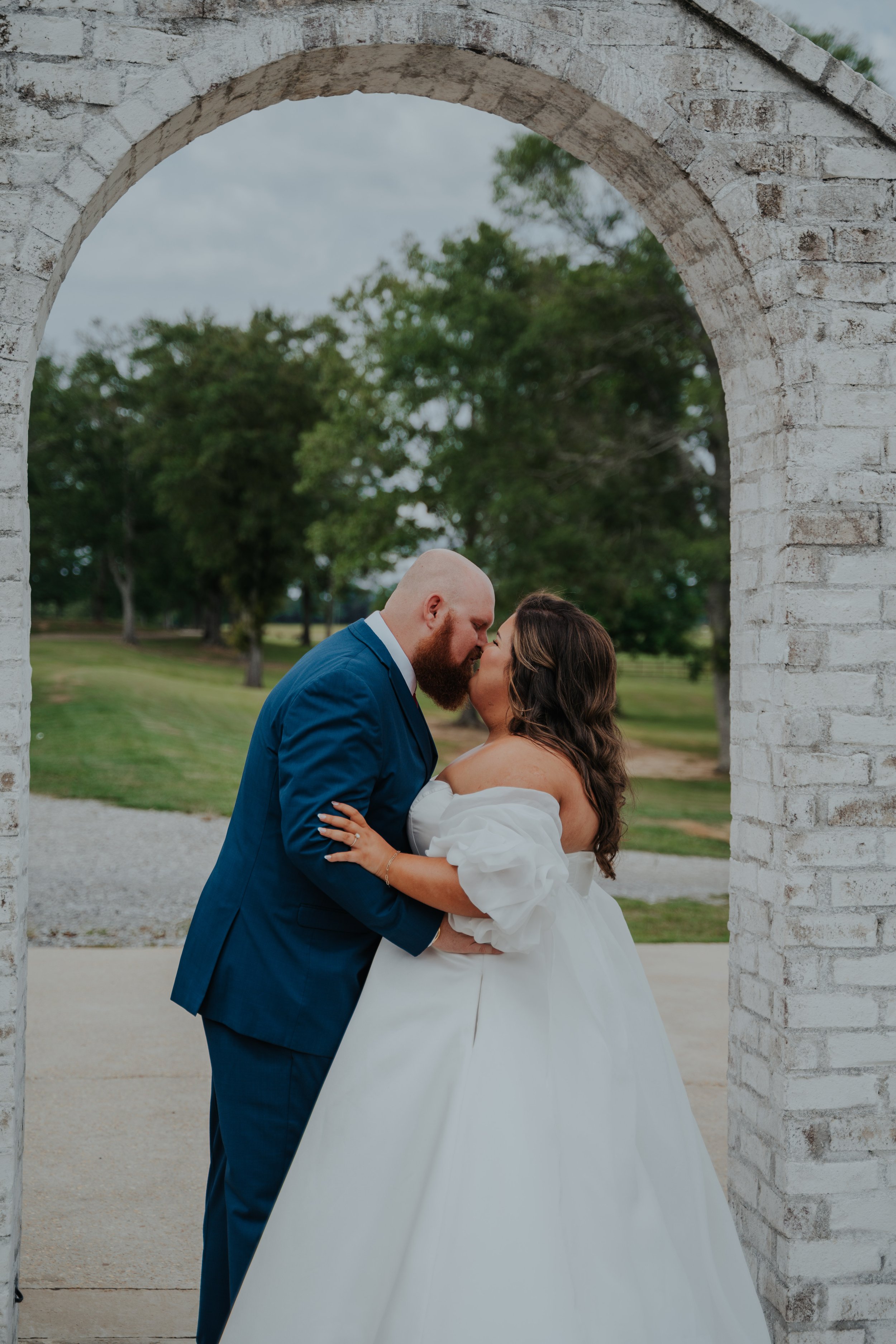 A bride and groom share a kiss under a white brick archway outdoors, with green trees and gray sky in the background.