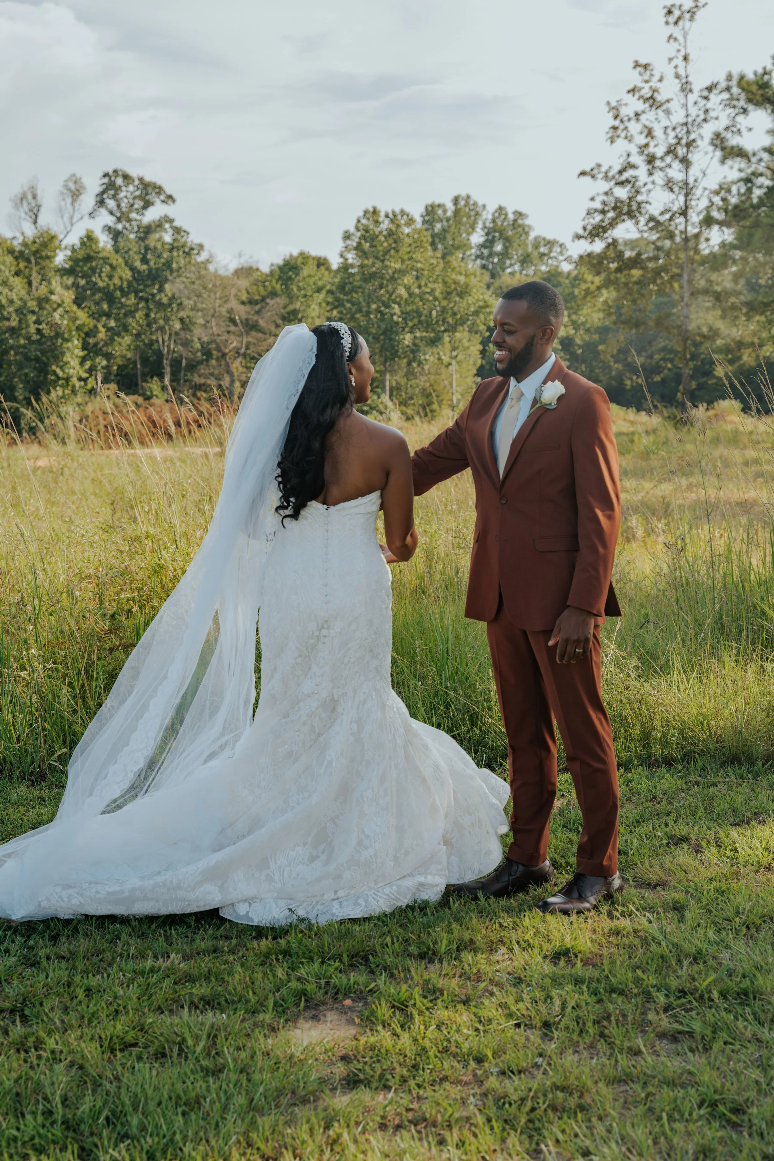 A bride and groom holding hands outdoors in a grassy field, facing each other and smiling, with trees and a cloudy sky in the background.