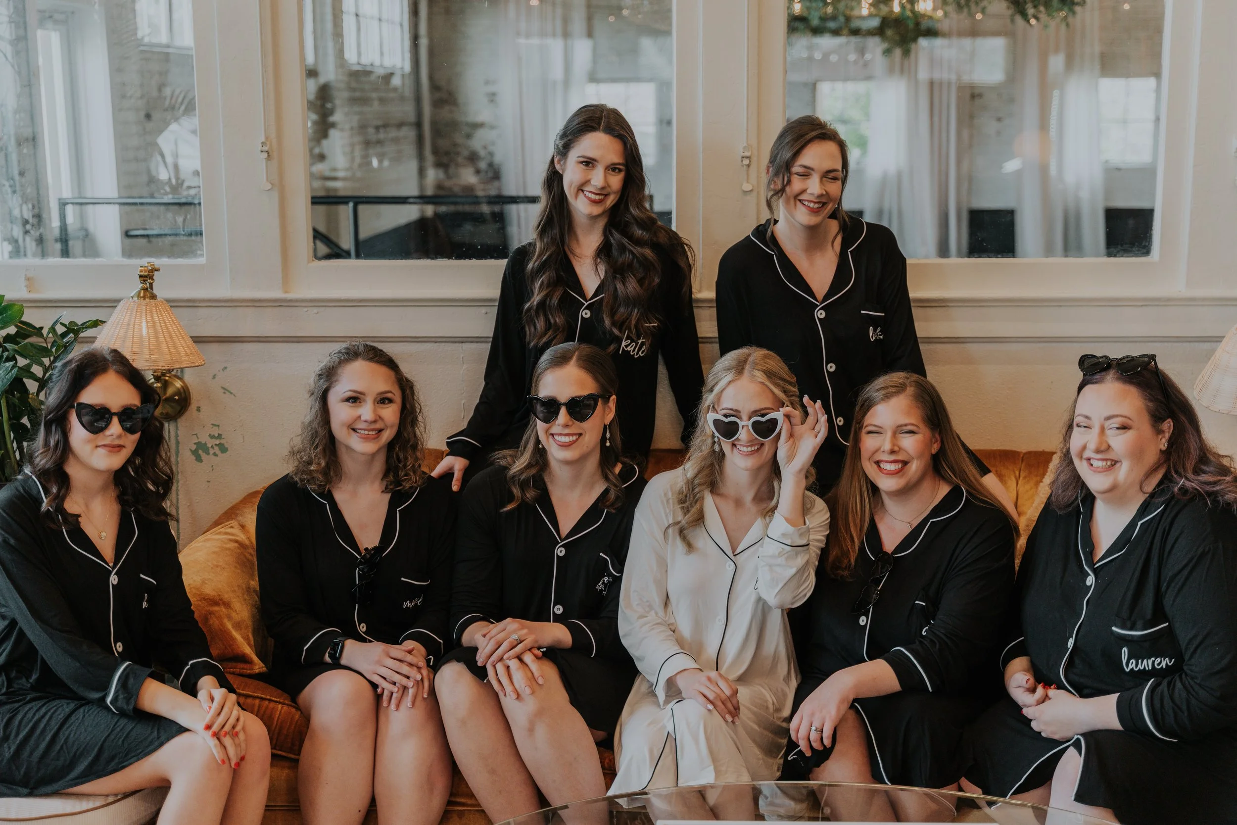 Eight women sitting and standing in pajamas, smiling in a cozy room with white walls, a sofa, and large windows. Some women wear sunglasses, and one woman has a white outfit and is adjusting her glasses.