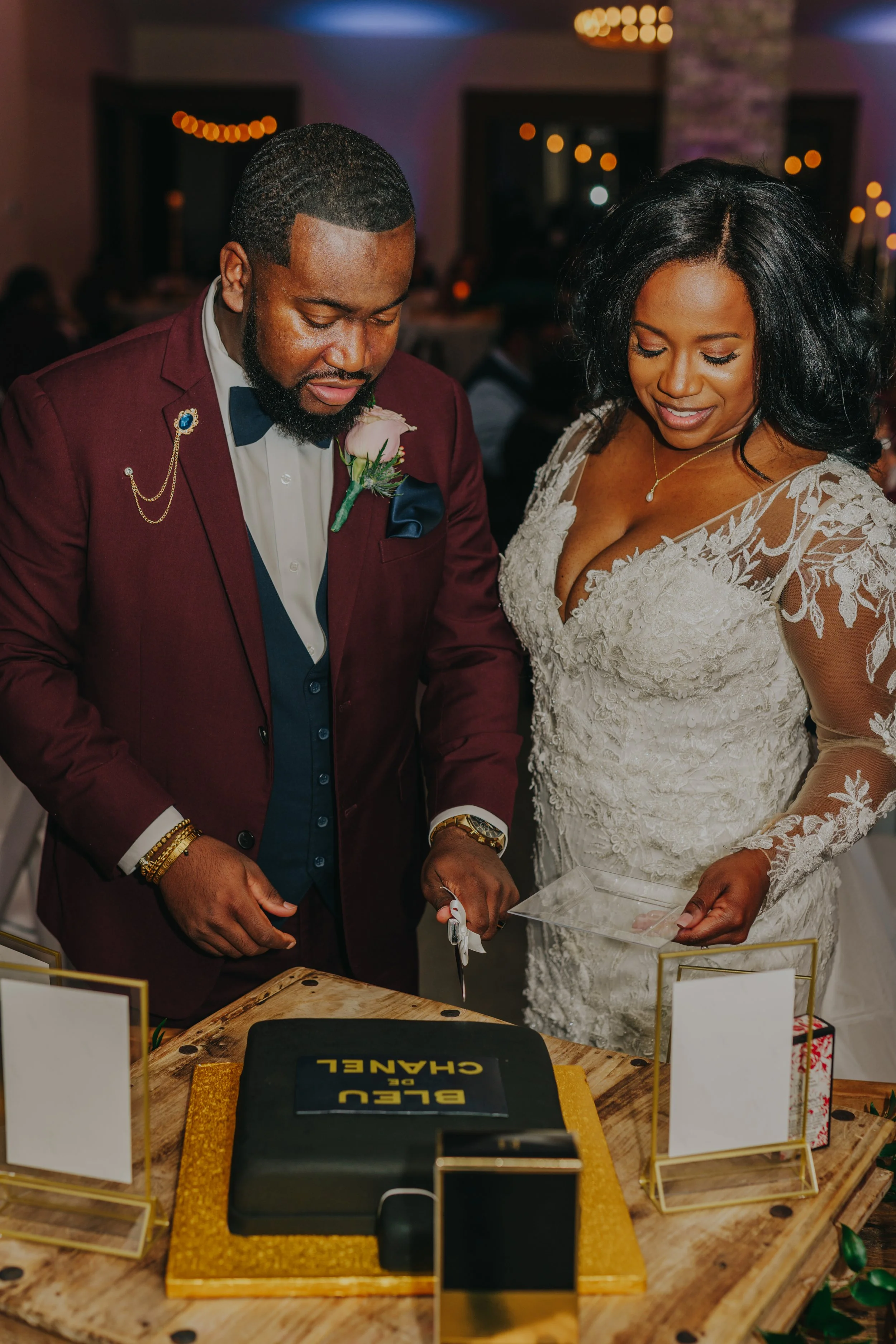 A couple at their wedding cutting a cake together. The groom is wearing a burgundy suit with a boutonniere, and the bride is in a lace wedding dress. They are smiling and looking down at the cake.
