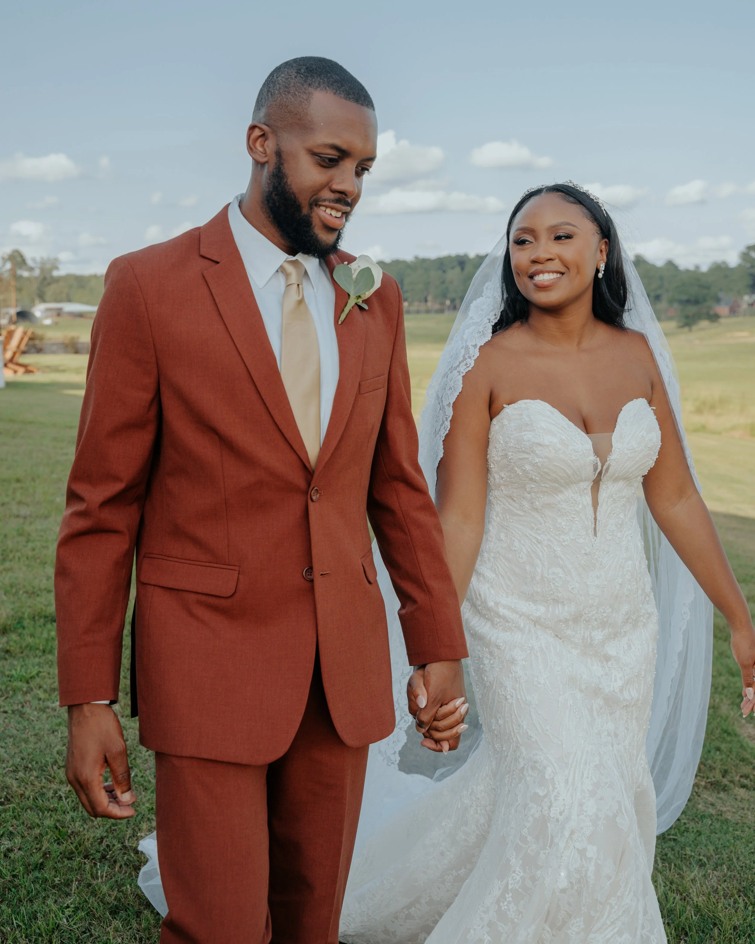 A newlywed couple walking outdoors on a sunny day, holding hands, with a grassy field and blue sky with clouds in the background.