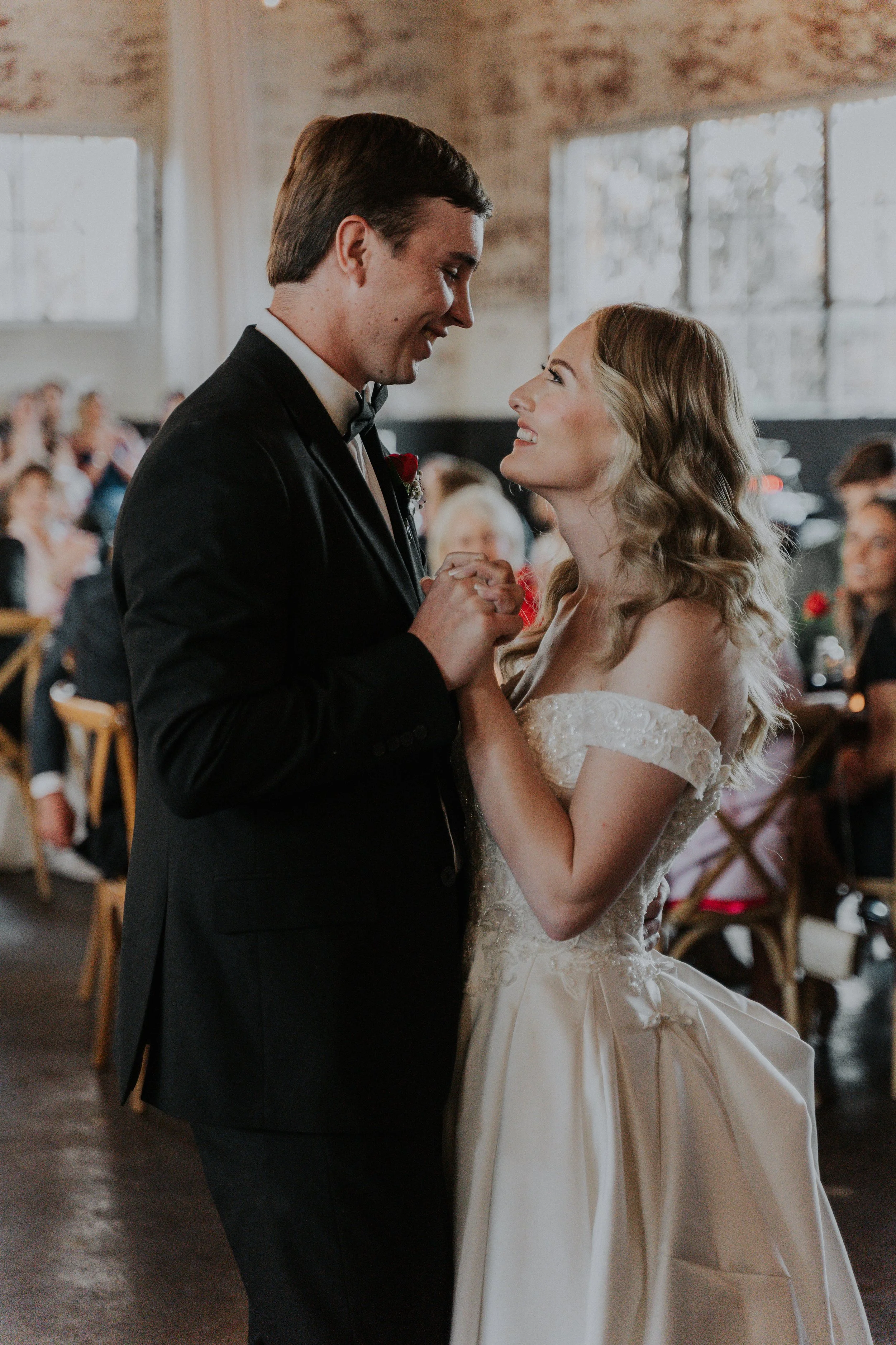A bride and groom share a dance at their wedding reception, holding hands and smiling at each other.