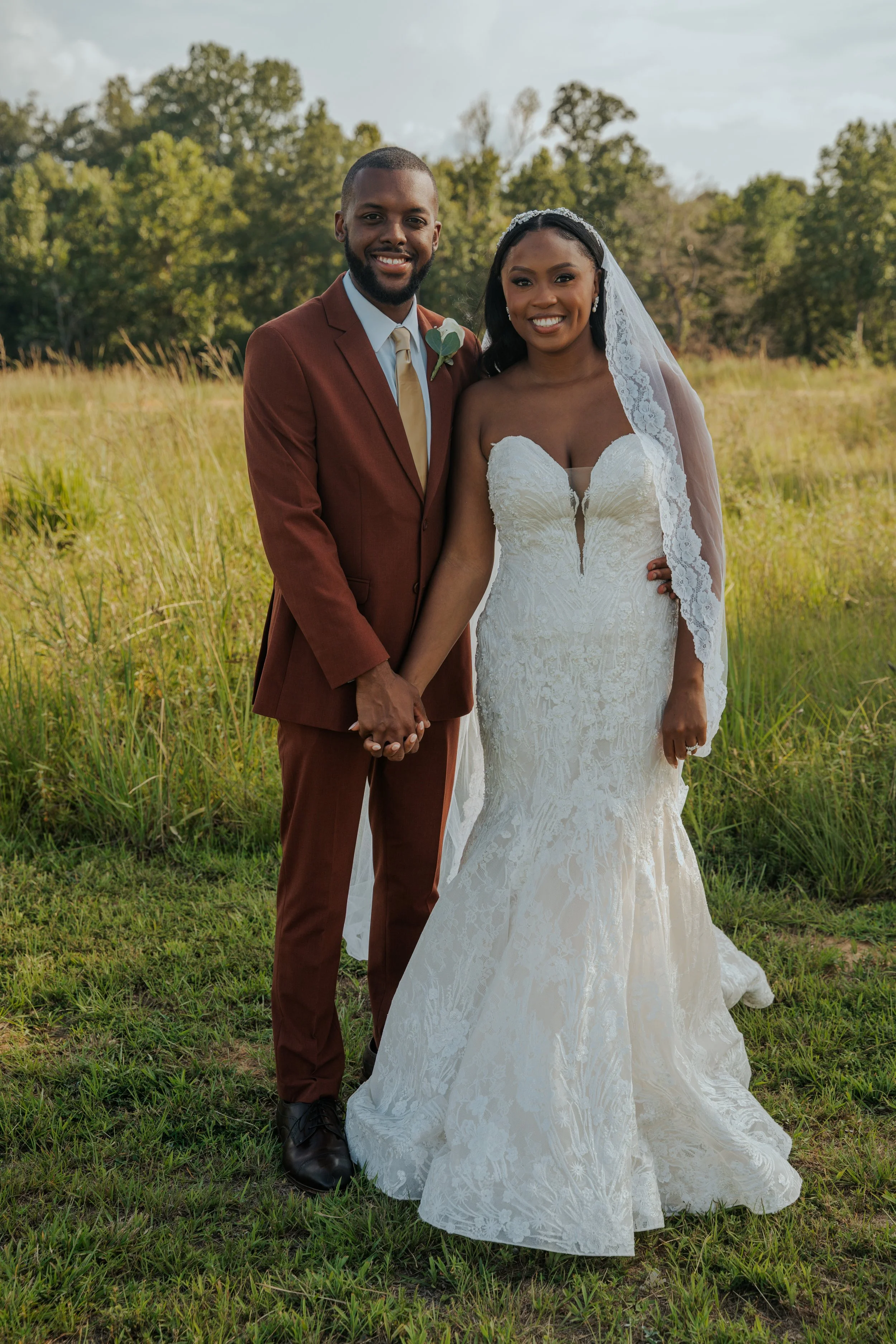 A newlywed couple holding hands outdoors in a grassy field, smiling at the camera, during daytime.
